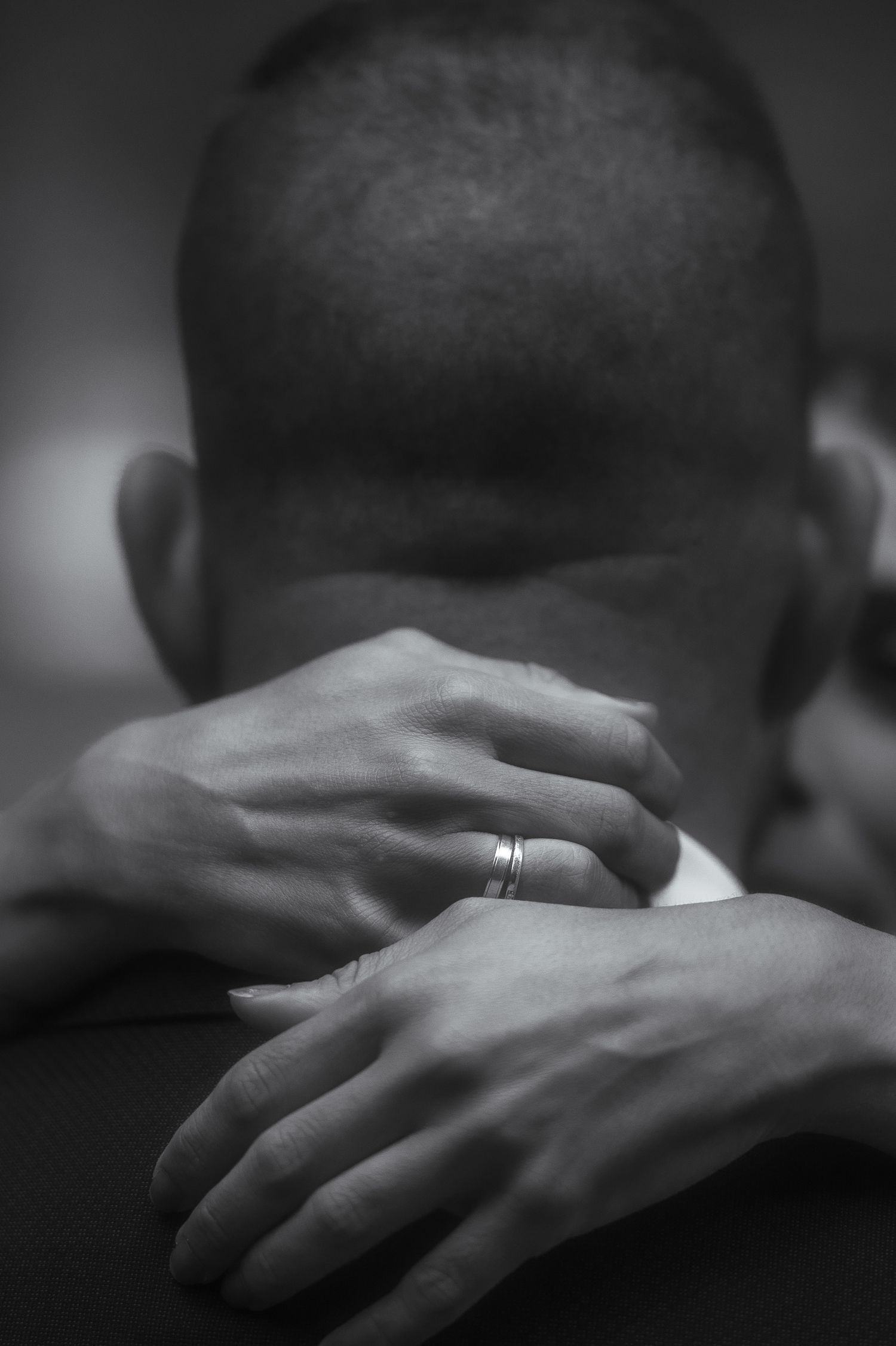 Black and white close-up image of hands with a wedding ring against a dark background.
