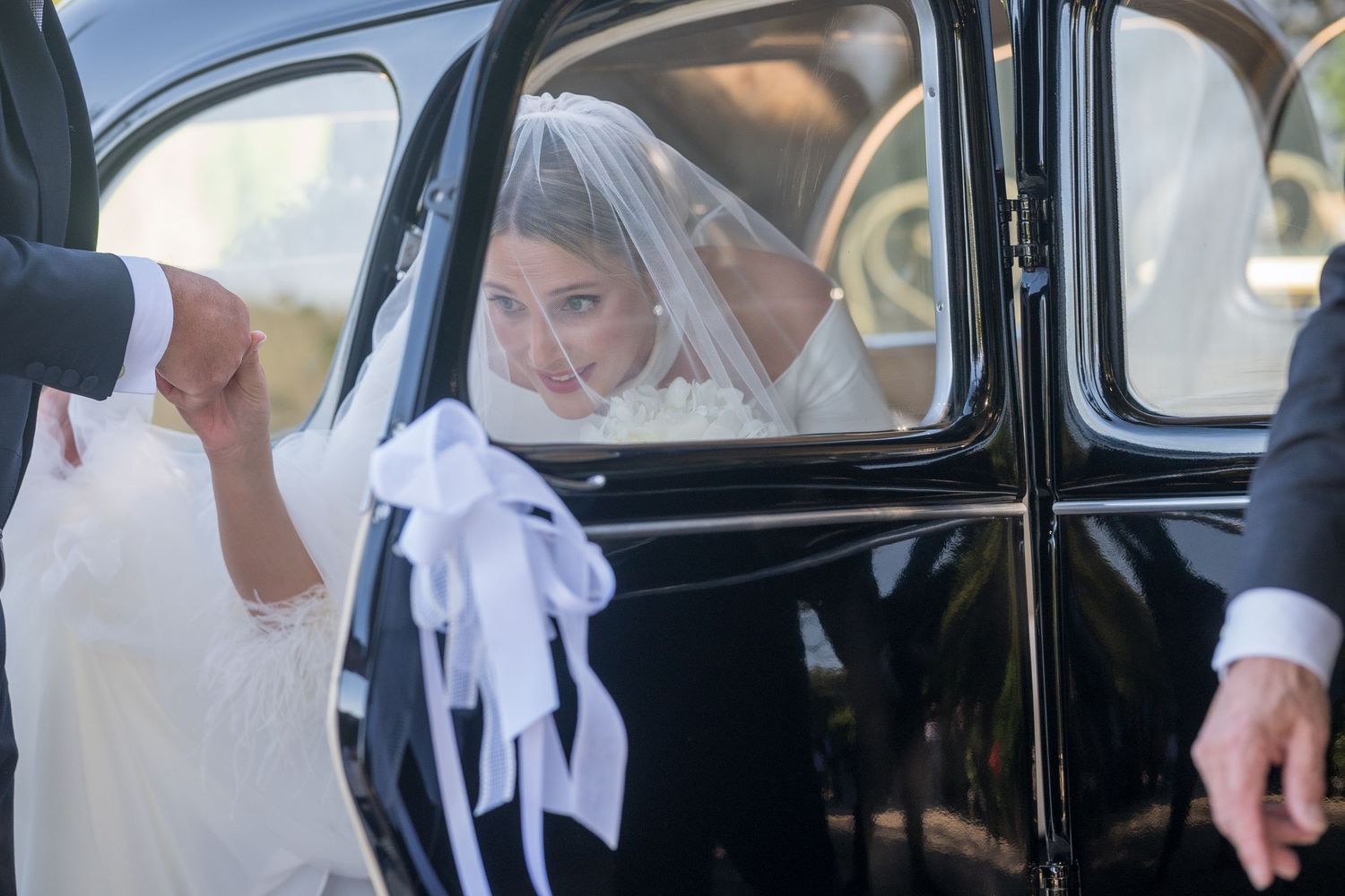 A person in a white veil looks out from a vintage black car window on their wedding day.