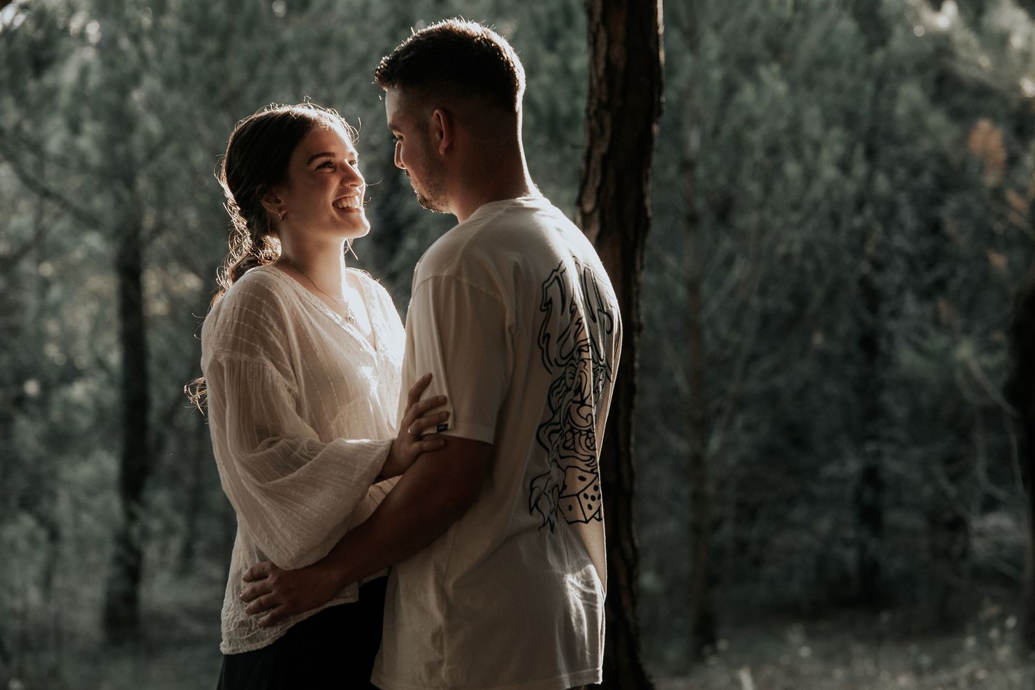 A couple shares an intimate moment in a moody forest setting with dramatic lighting and shadows.