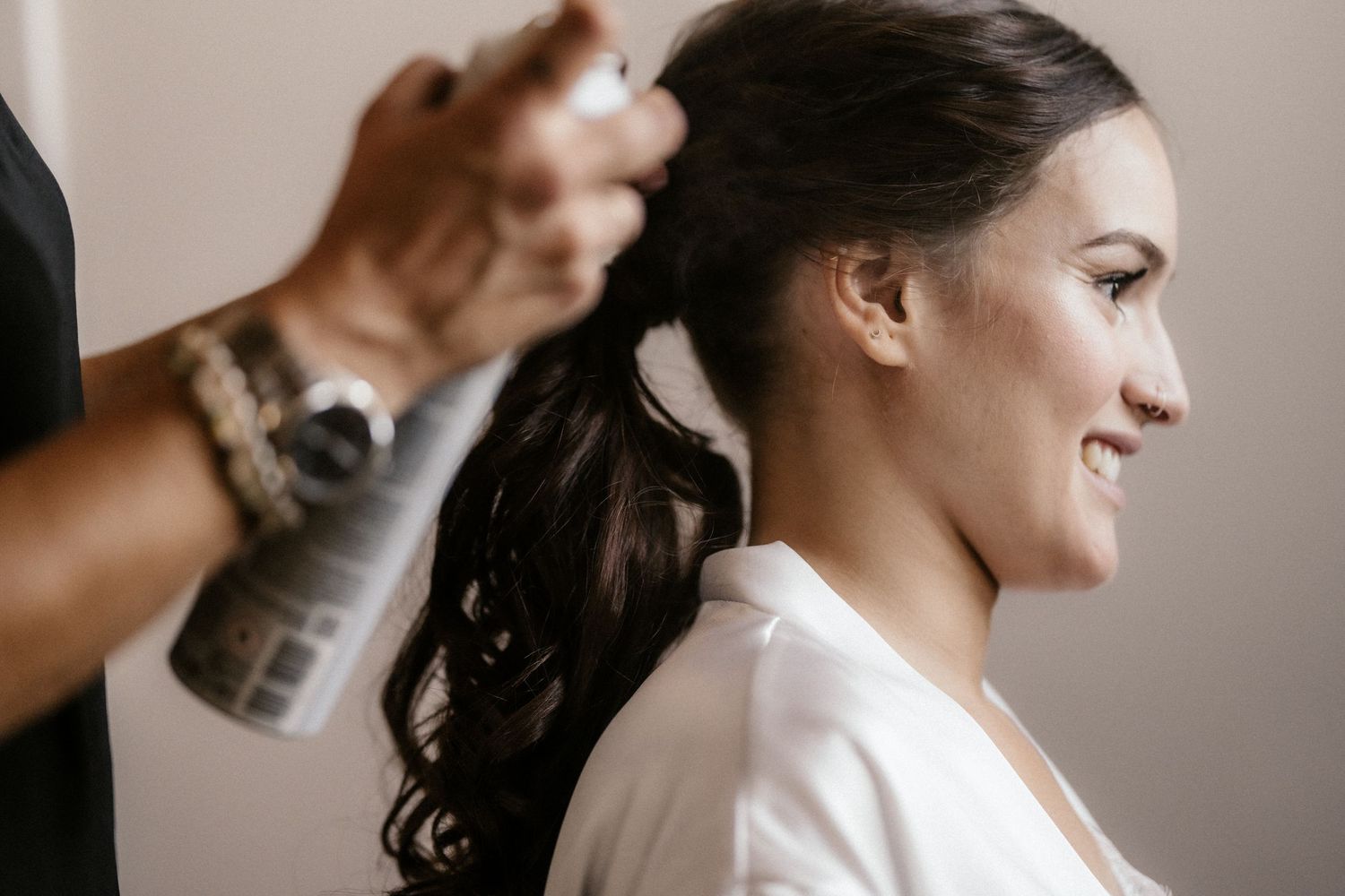 Hairstylist spraying hairspray on dark-haired woman's styled updo in salon.