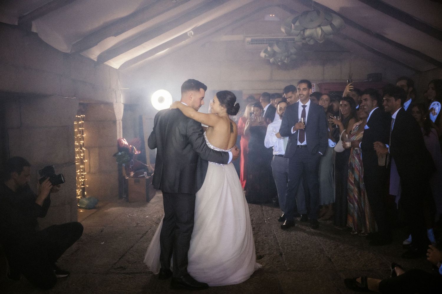 A wedding couple shares their first dance surrounded by guests in a dimly lit reception venue with fog effects.