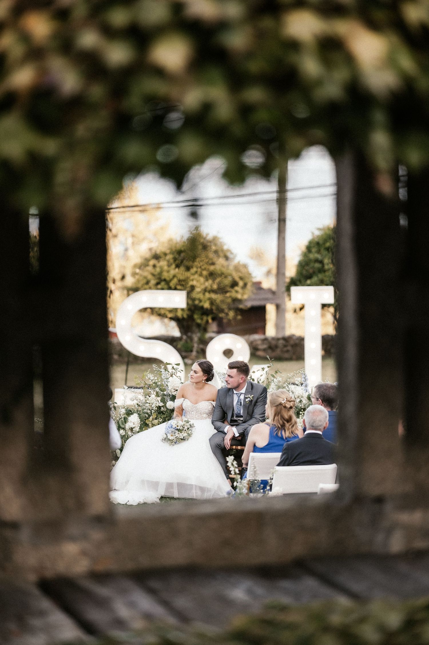 A wedding ceremony viewed through a rustic window frame with guests seated outdoors.