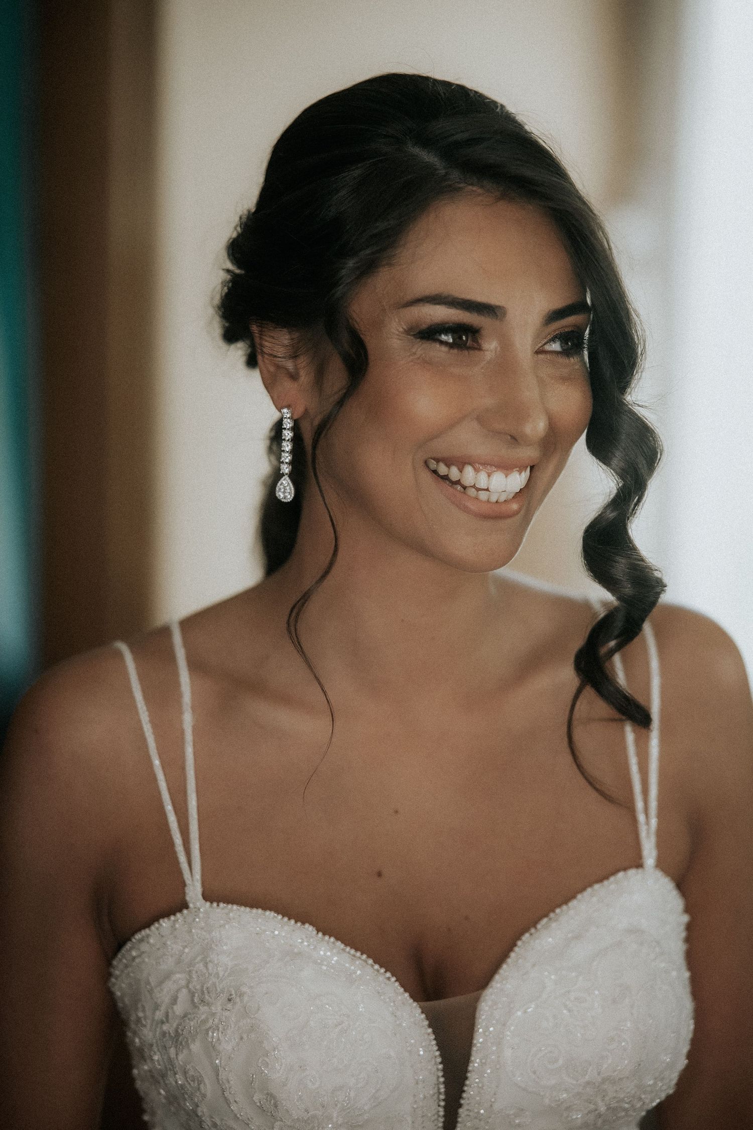 A bride in a white strapless wedding dress smiles radiantly while getting ready for her wedding ceremony.