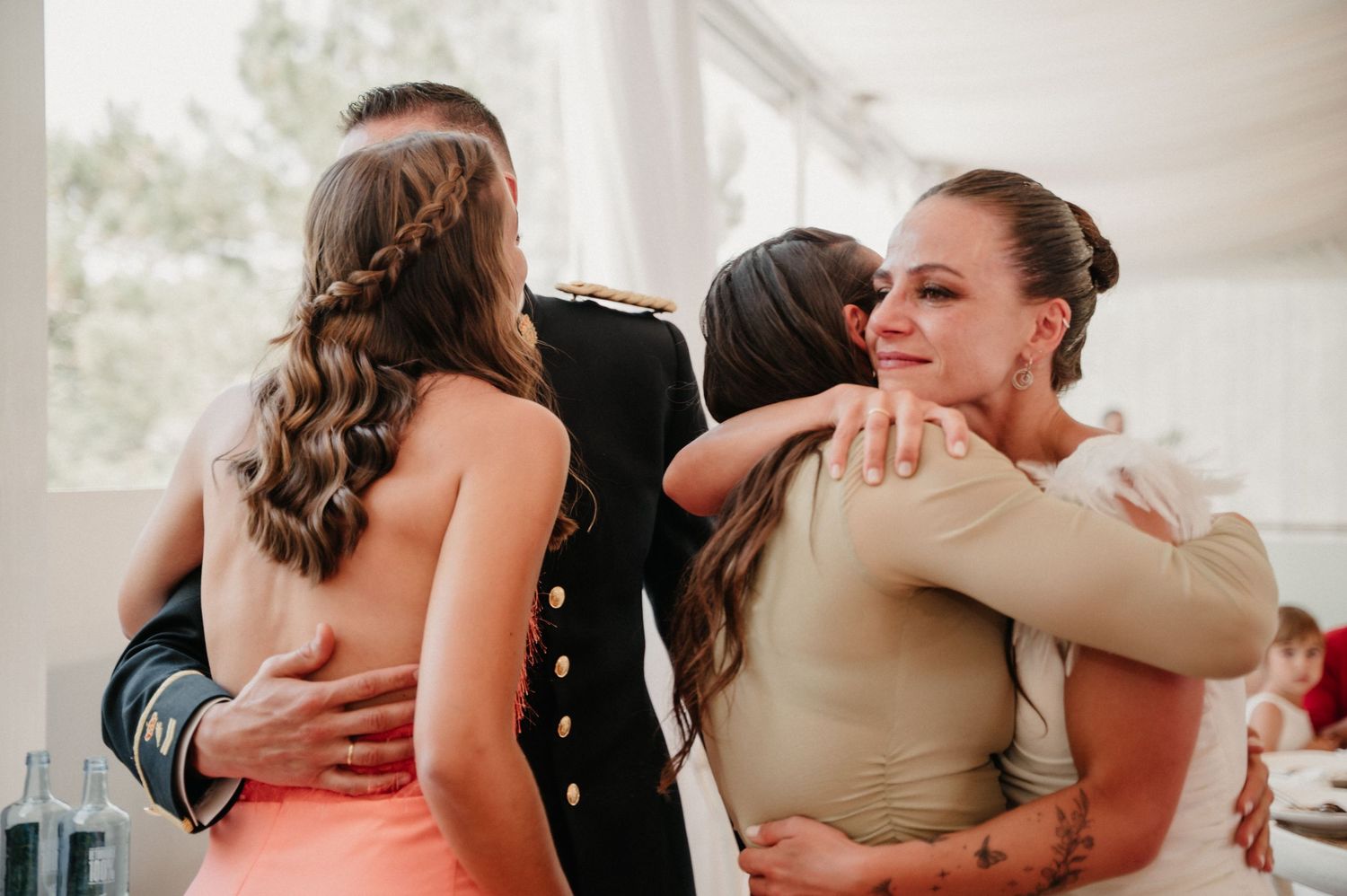 Emotional wedding guests embrace during reception celebration in bright indoor venue.