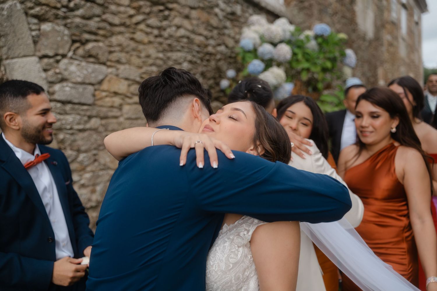 Wedding party celebrates with guests in front of a rustic stone wall backdrop.