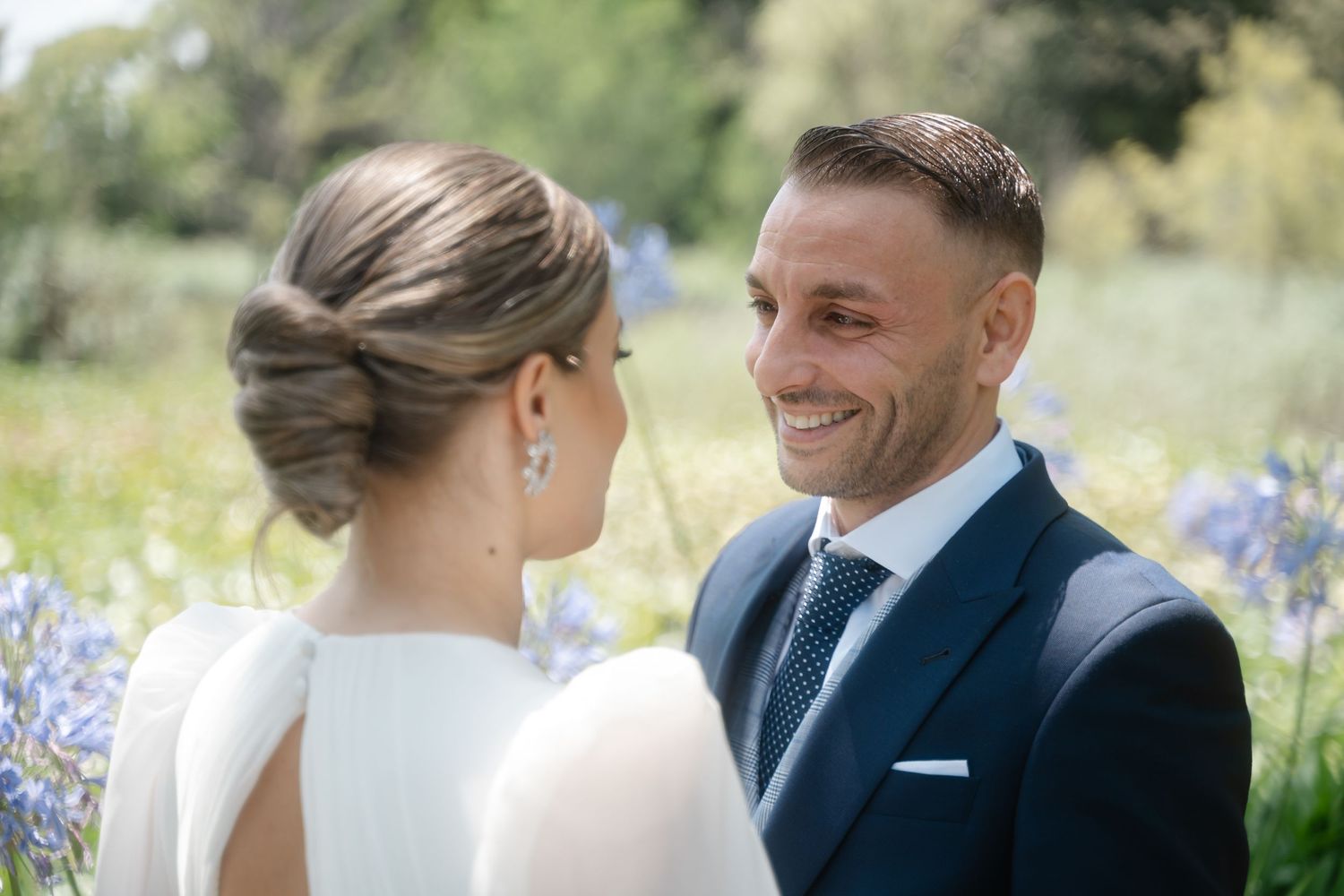 Happy couple shares intimate moments during outdoor wedding photoshoot with purple flowers in background.