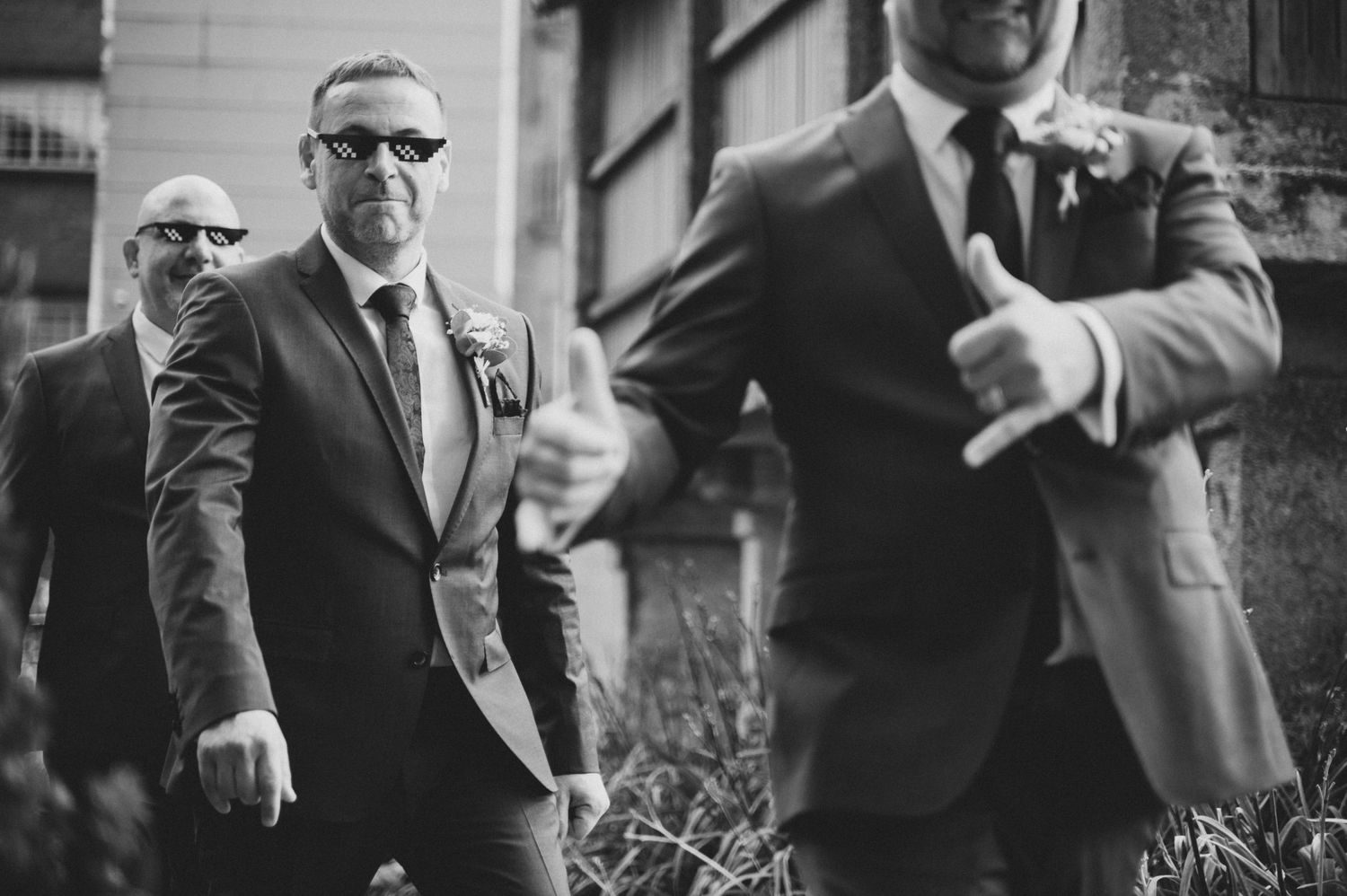 Black and white photo of groomsmen in dark suits and sunglasses walking together before a wedding ceremony.