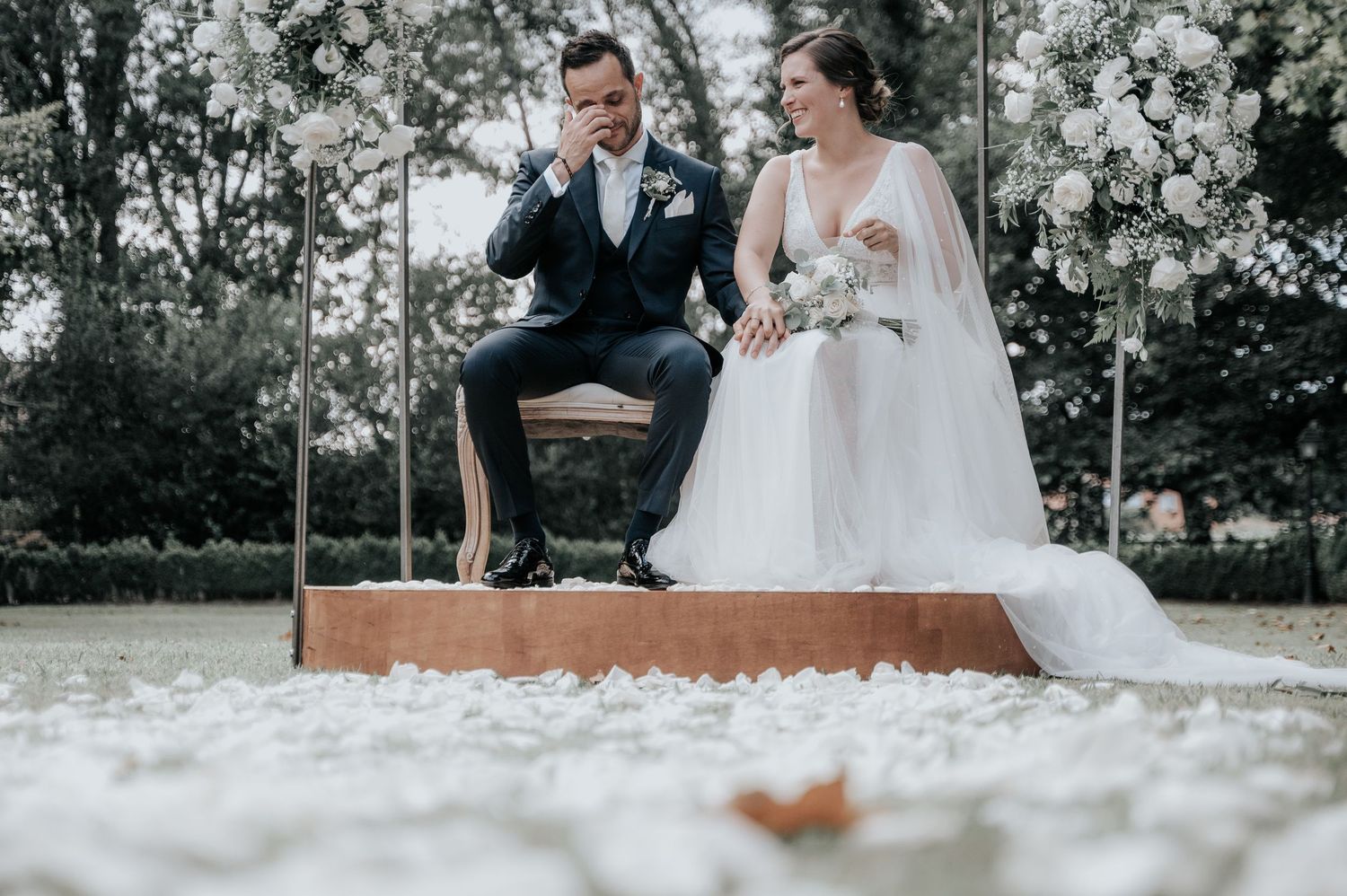 A couple sits together on a rustic wooden bench surrounded by white floral decorations during an outdoor wedding ceremony.