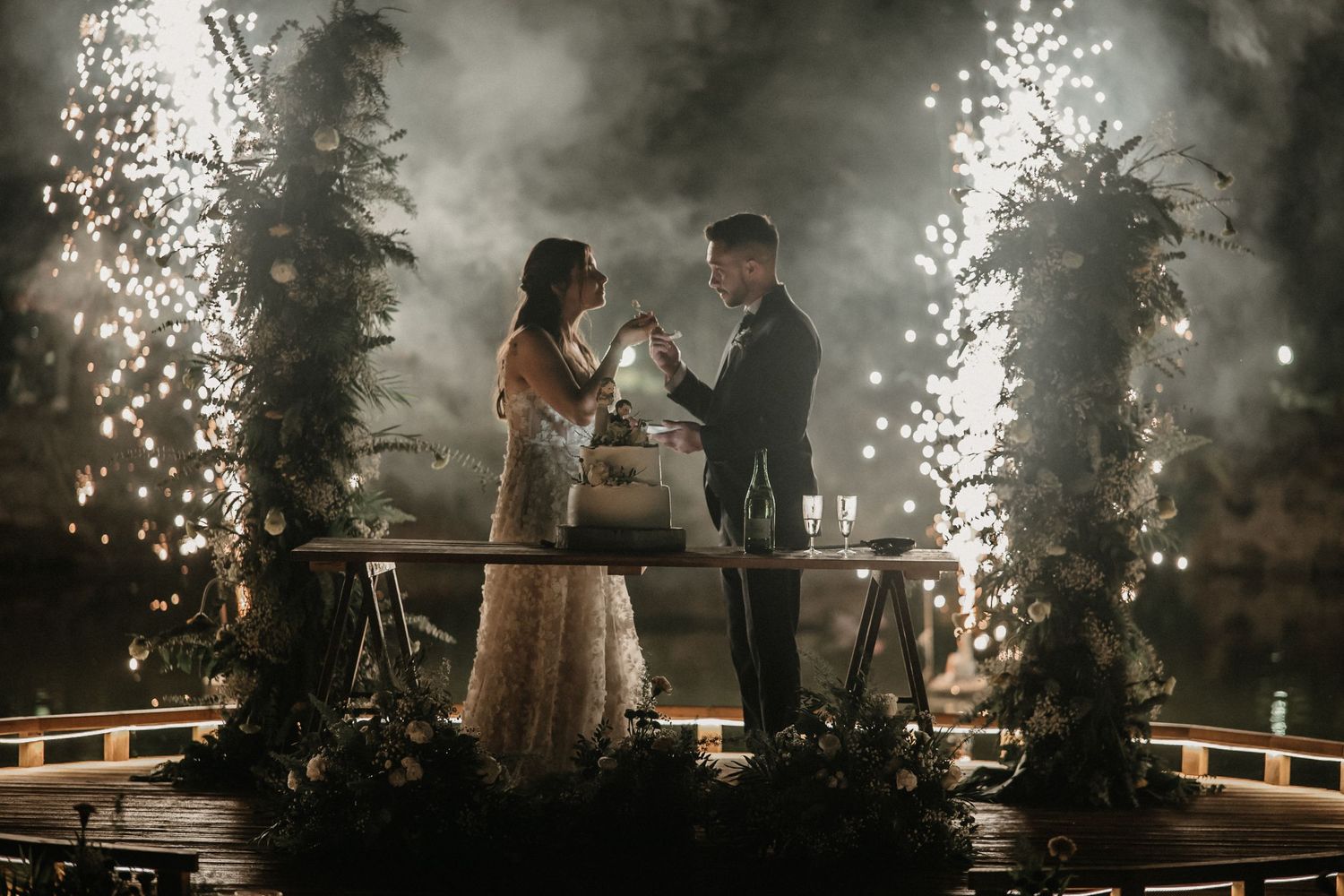 Silhouetted figures stand on a misty wooden deck surrounded by sparklers and atmospheric fog at night.