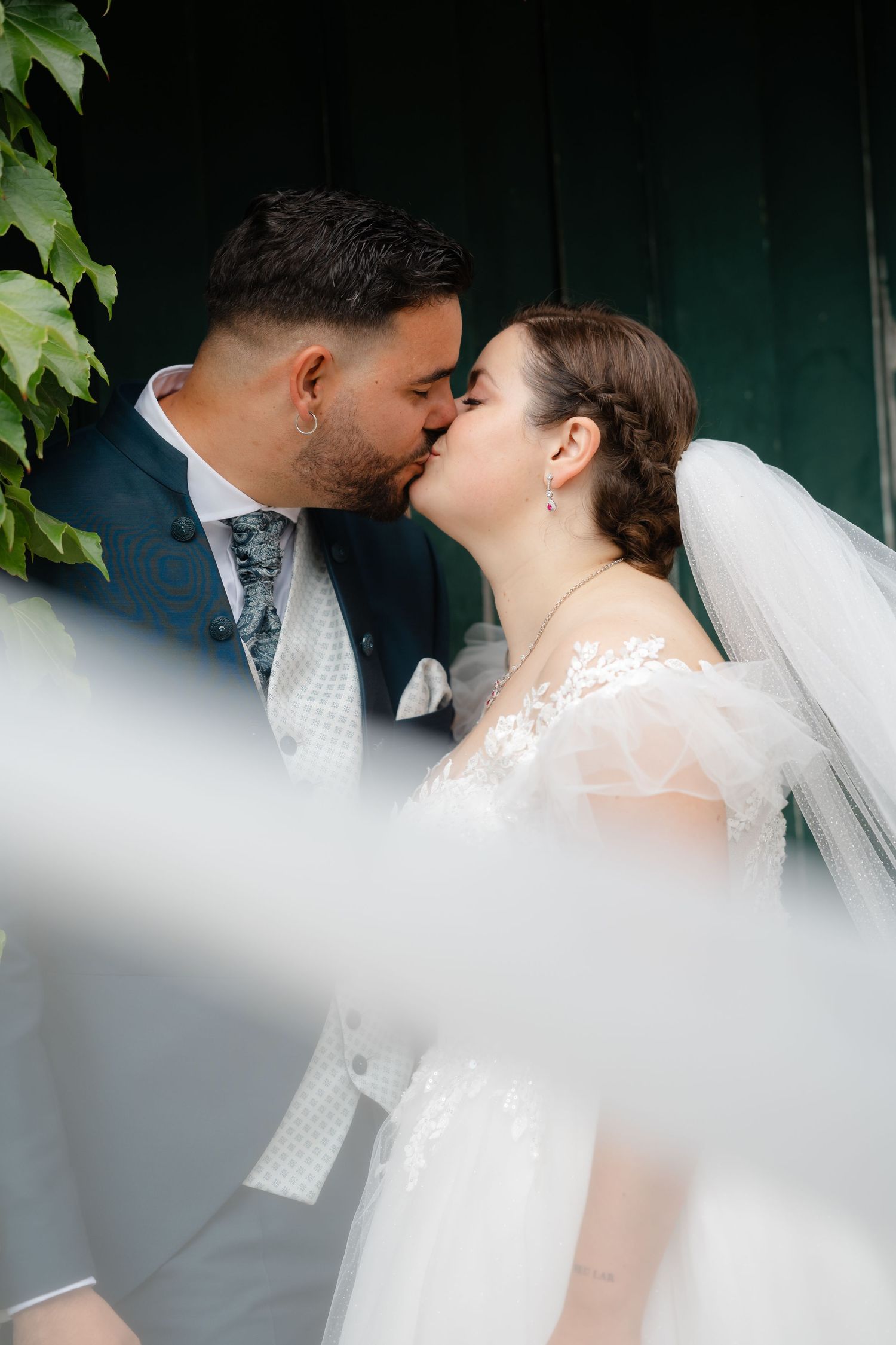 A romantic wedding couple shares a tender kiss against a green backdrop with soft lighting.