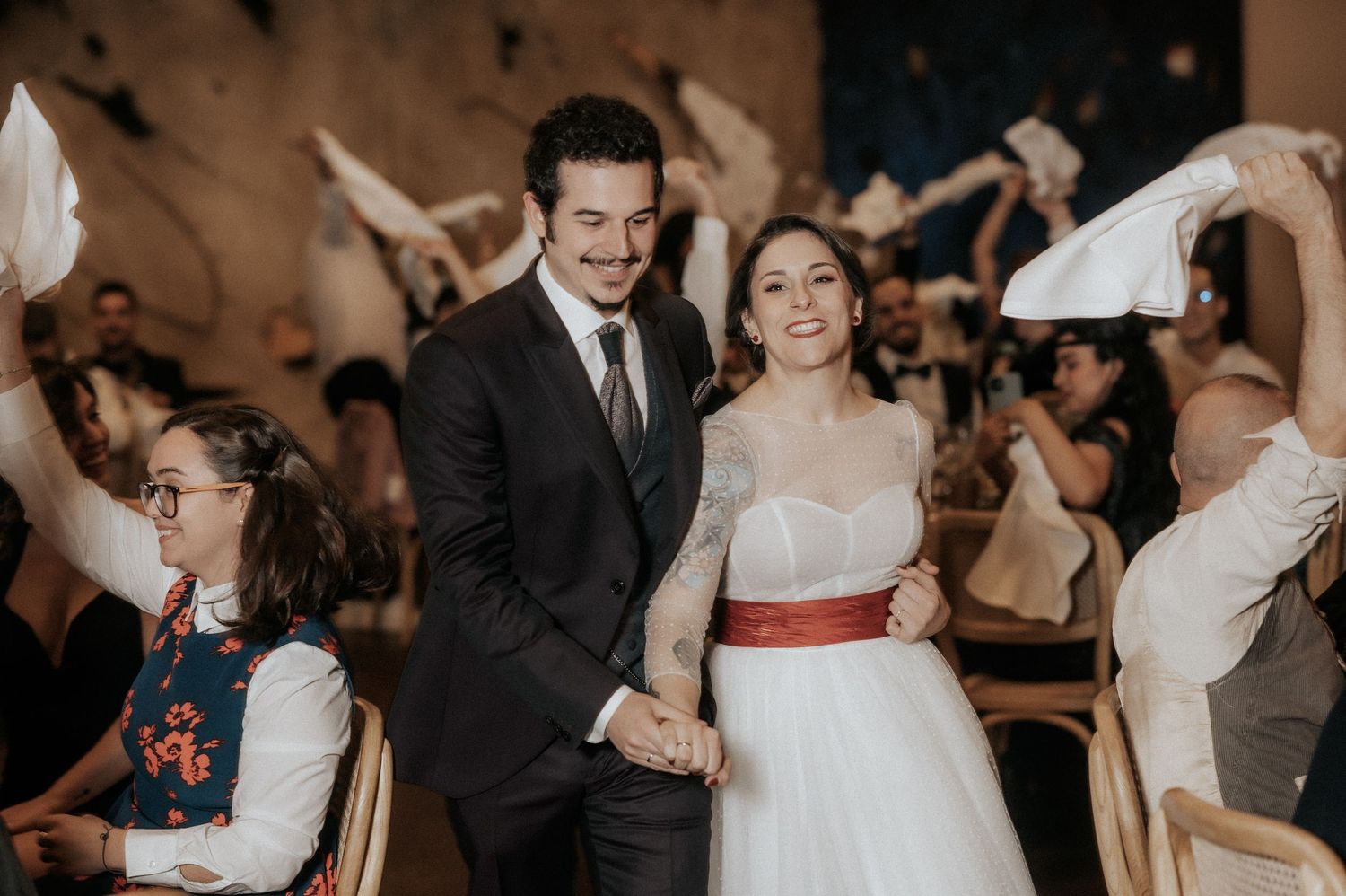 A couple dances together at their wedding reception while guests celebrate around them in a dimly lit venue.