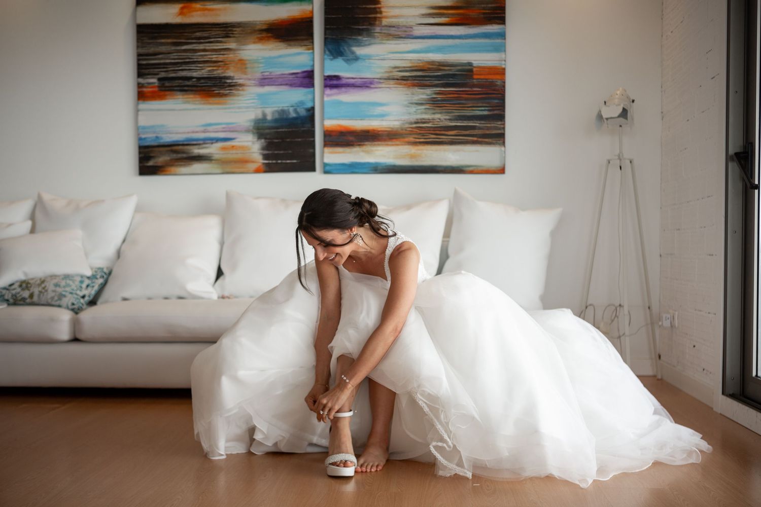 A bride in a flowing white wedding dress and heels walks across a room with sunset paintings on the wall.