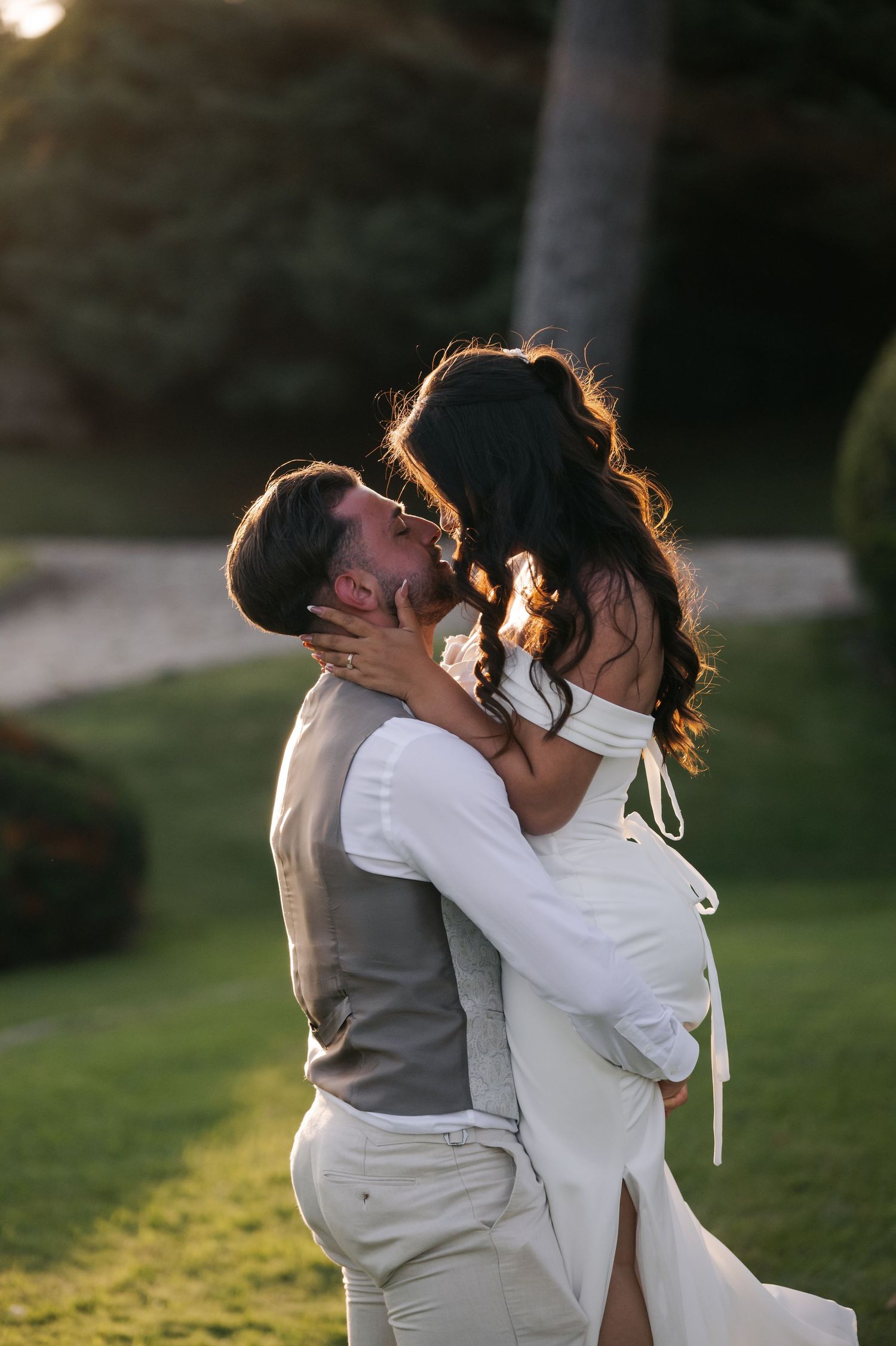 A romantic couple in gray and white attire share a passionate kiss at sunset in a grassy outdoor setting.