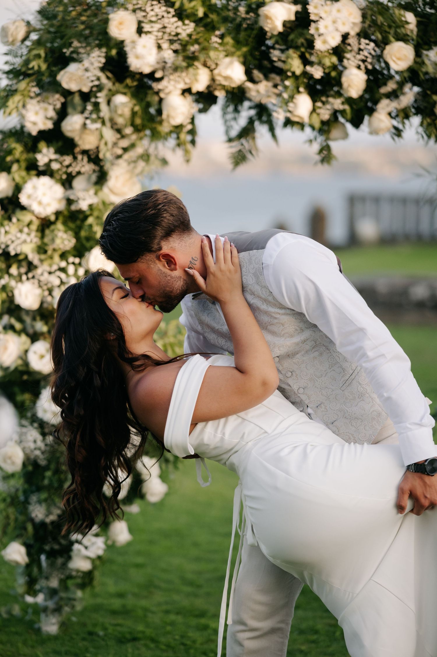 Romantic wedding couple sharing a dip kiss under a floral arch with white roses in an outdoor garden setting.