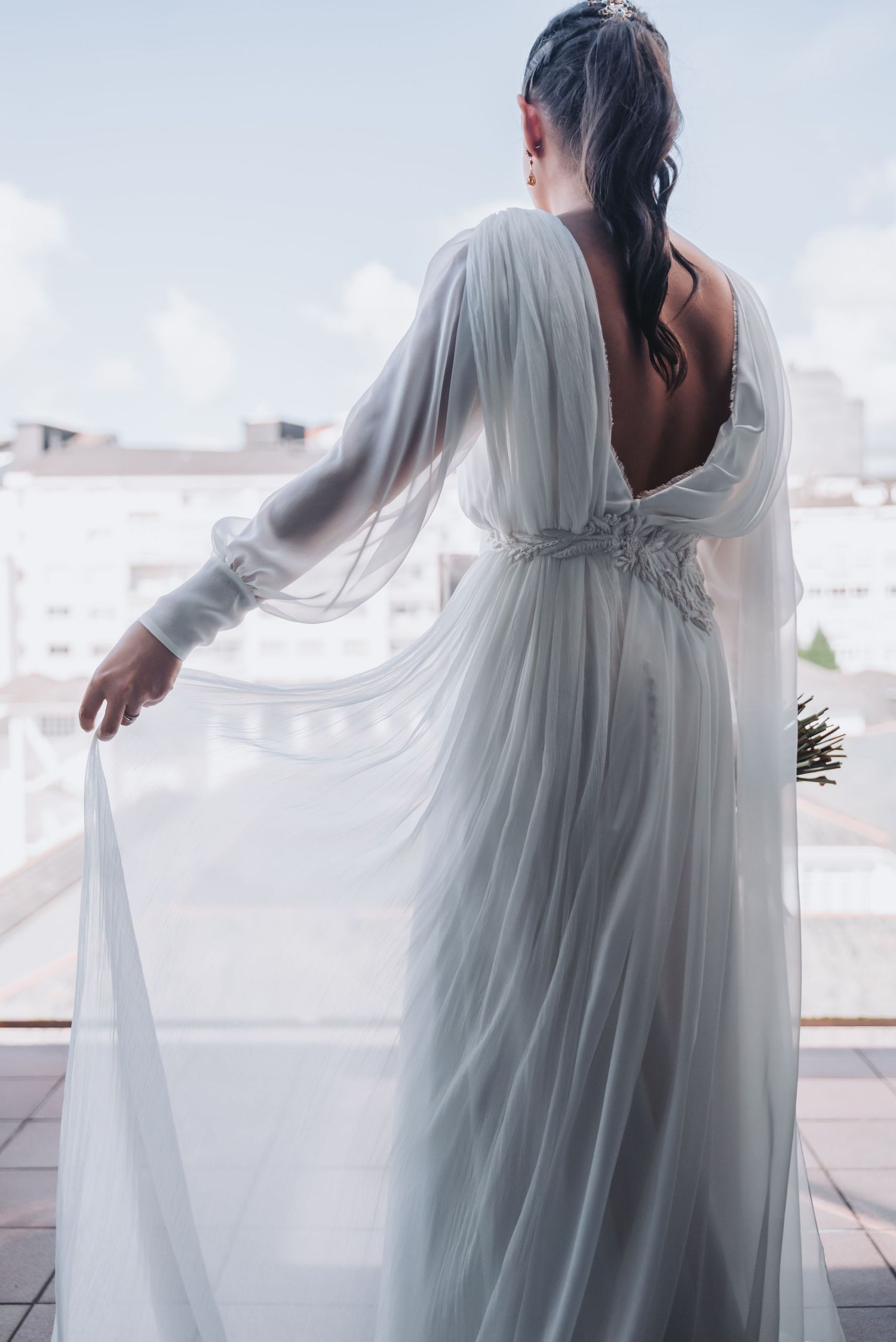 Back view of a flowing white wedding gown with sheer sleeves against an urban backdrop.