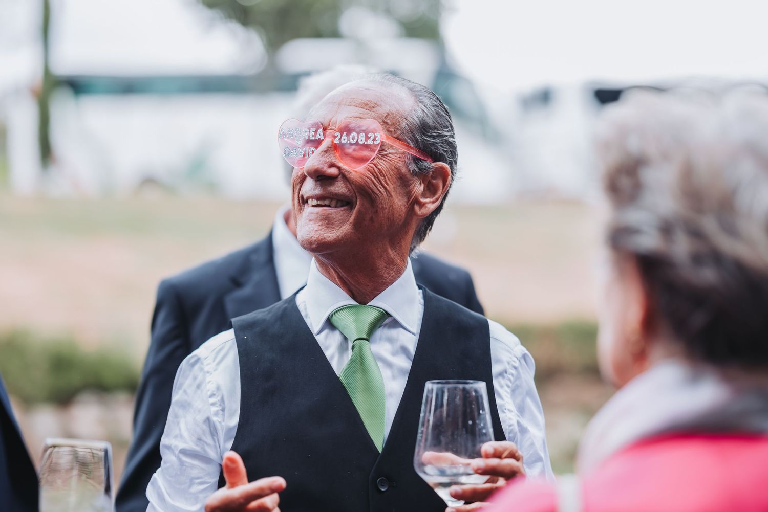 A person in a vest and green tie laughs heartily while holding a glass of red wine at an outdoor event.