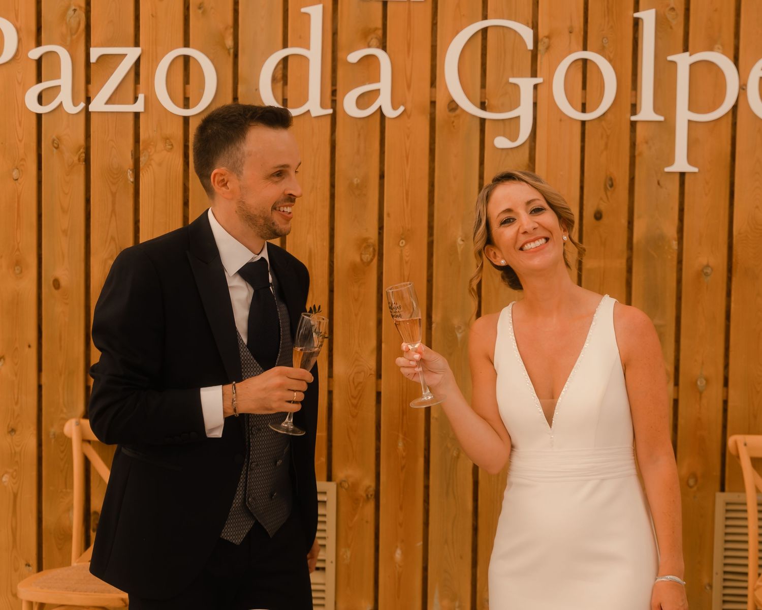 Wedding celebration scene with people toasting against a wooden paneled wall in warm lighting.