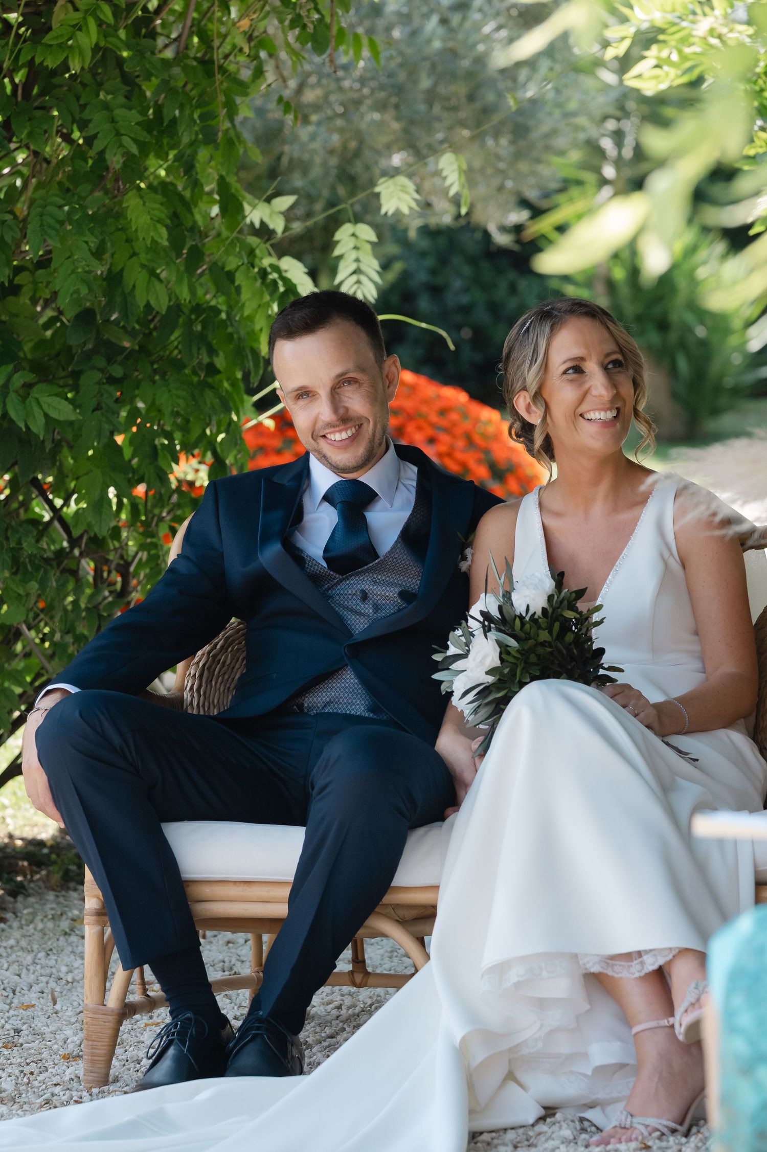 A couple in wedding attire share a joyful moment while sitting together on a wooden bench in a garden setting.