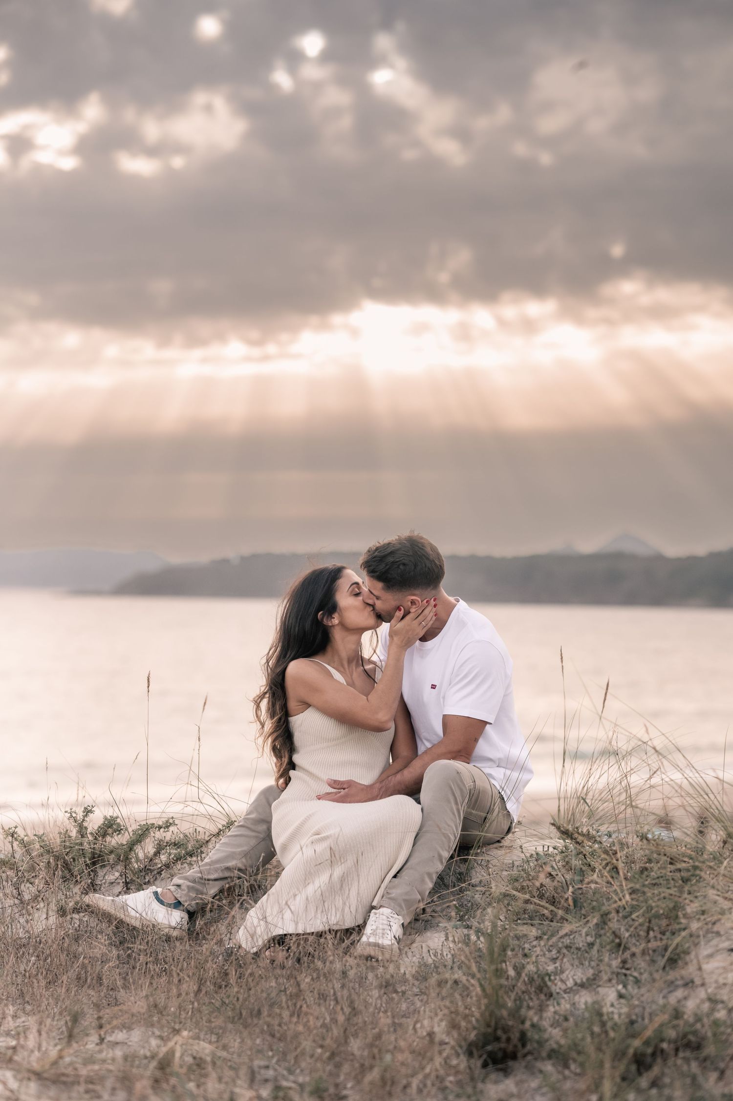 A couple shares tender moments while lying in tall grass during golden hour with mountains in the distance.