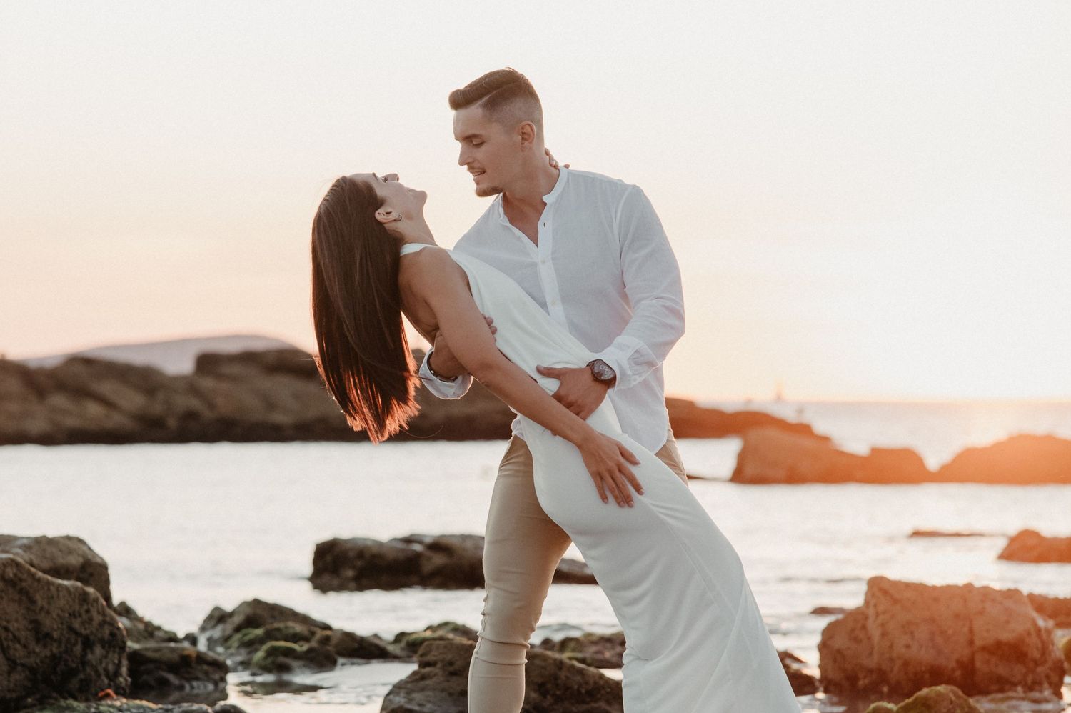 A romantic couple in white outfits embrace on a rocky beach at sunset with golden light illuminating the scene.