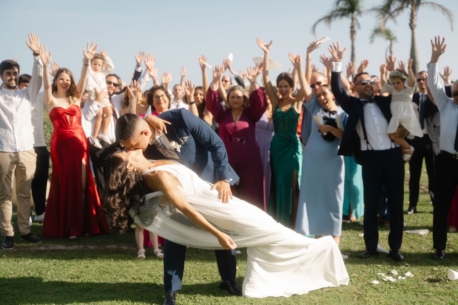 Wedding guests cheer and raise their arms as a couple celebrates during an outdoor ceremony with palm trees in view.