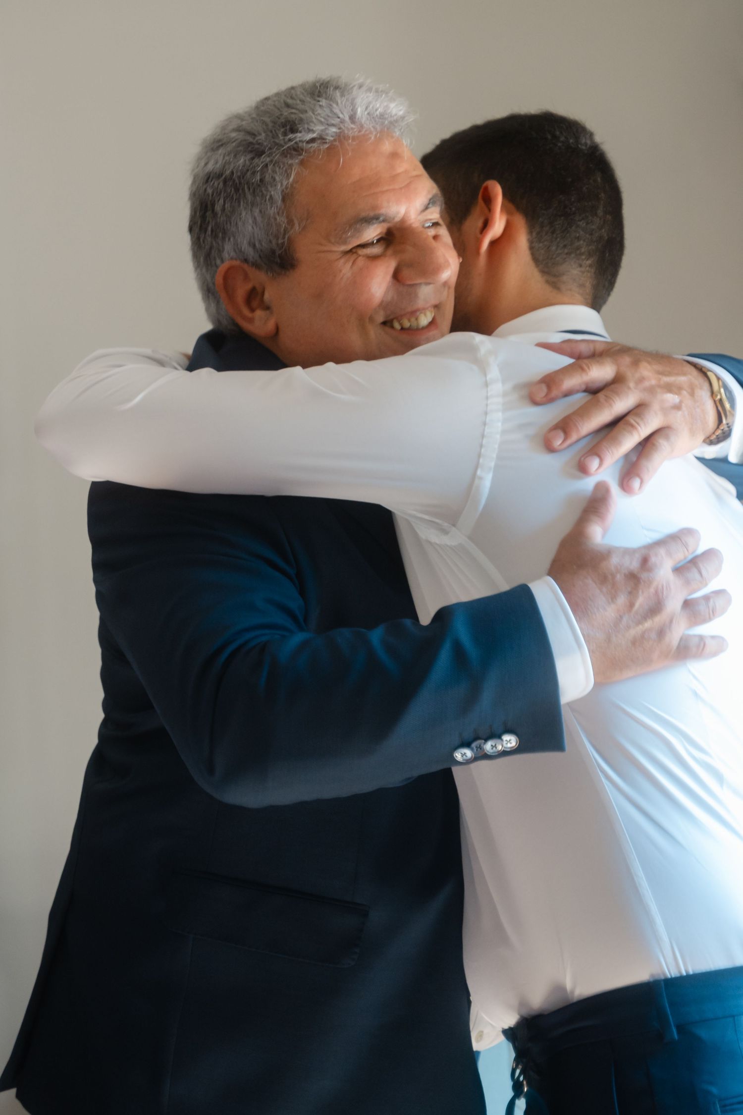Two people sharing a warm embrace in formal attire against neutral background.