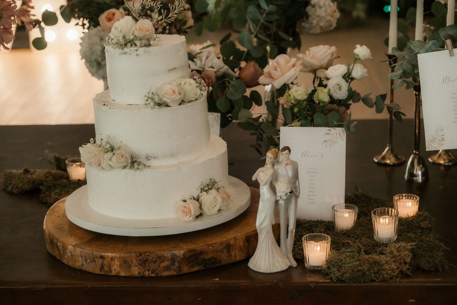 Elegant white wedding cake with floral decorations and candlelight on a rustic wooden base at a reception.