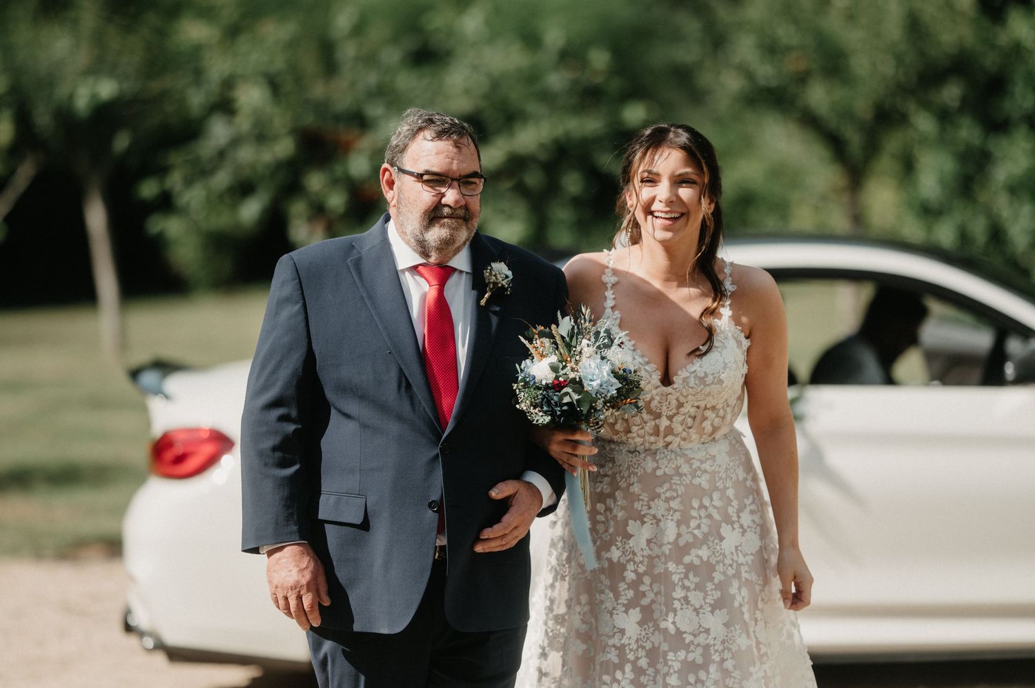 A couple in wedding attire walk together outdoors on a sunny day with trees in the background.