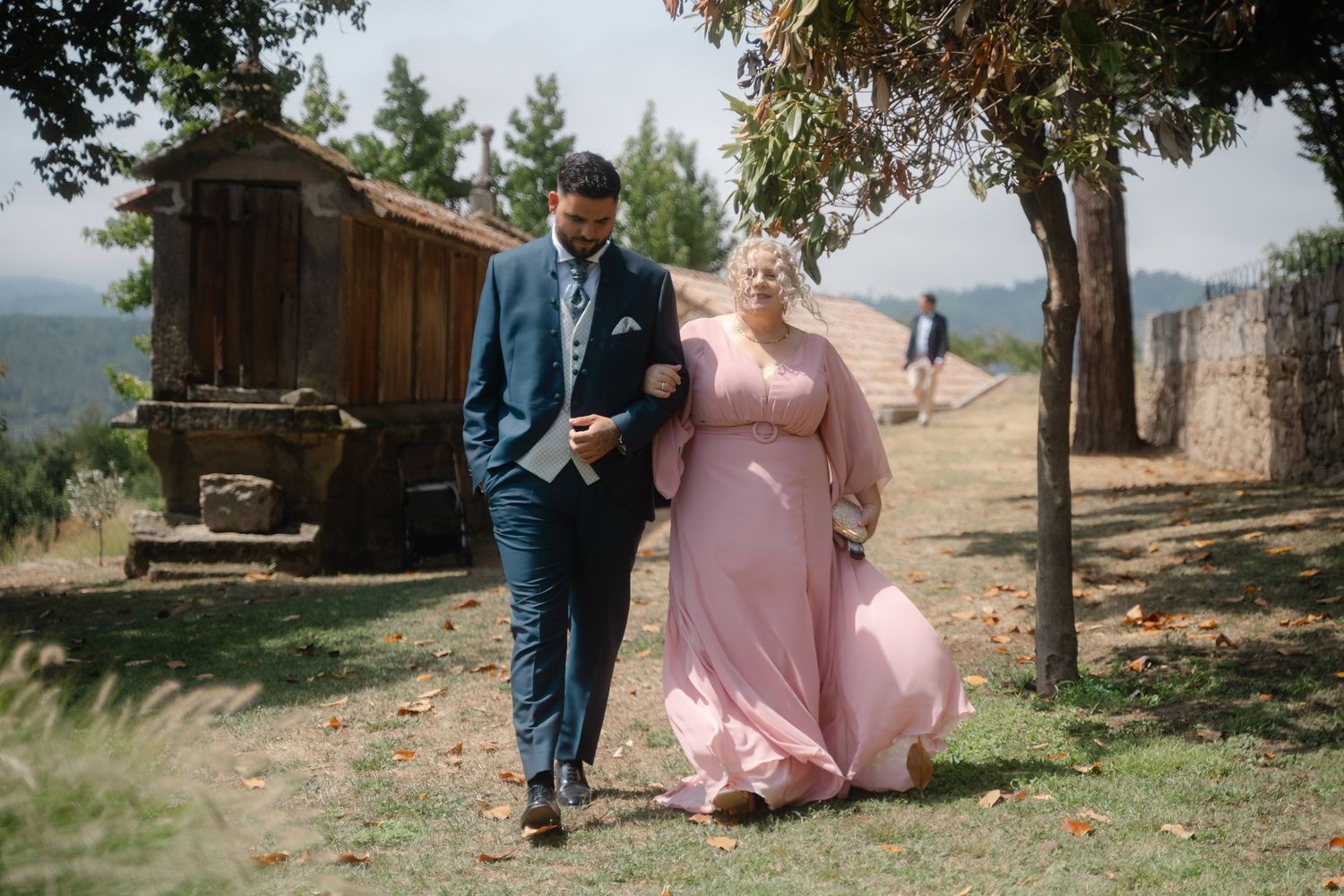 A couple in formal attire walks together through a scenic mountain landscape on an autumn day.