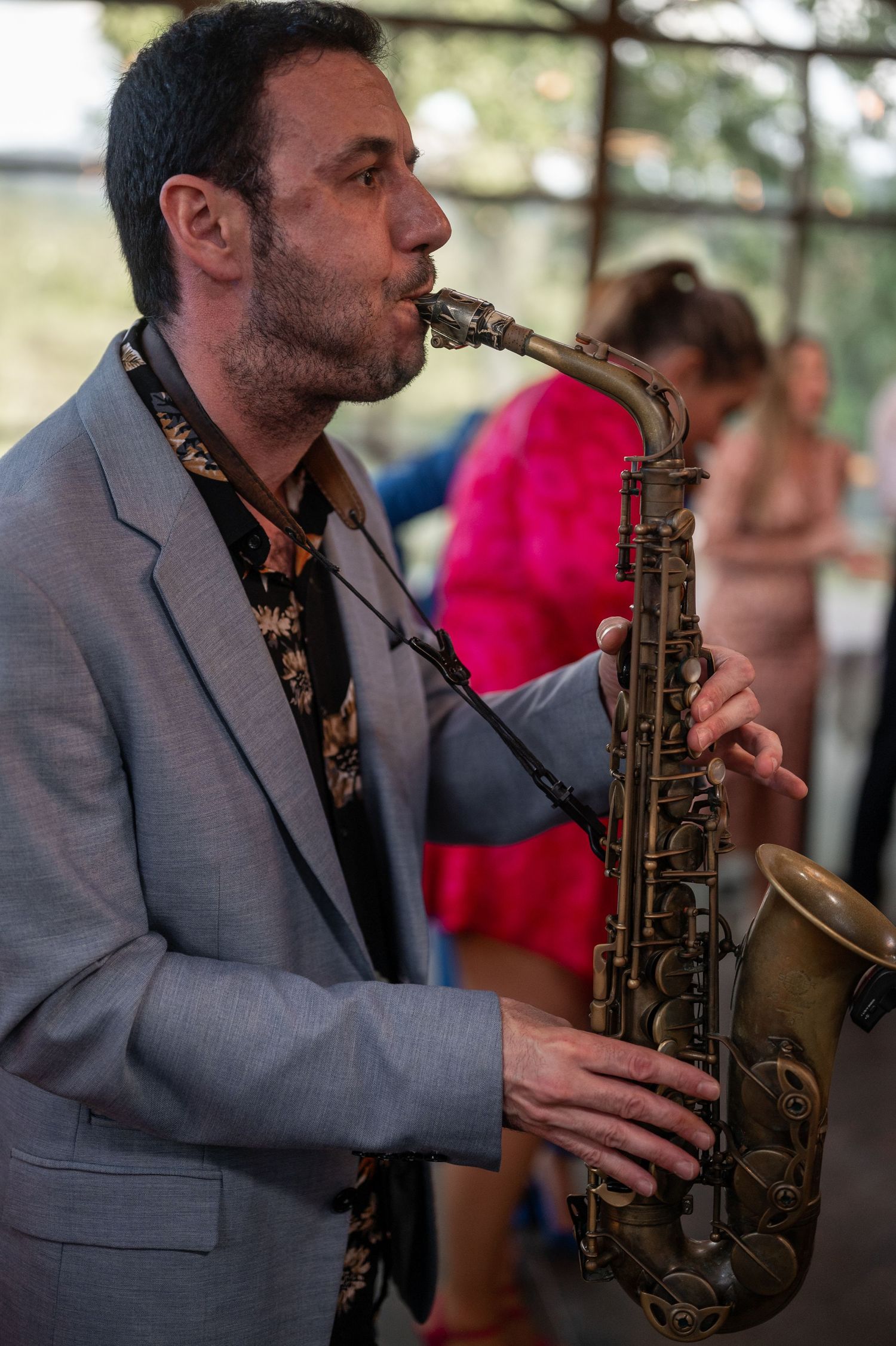 A musician in a gray suit jacket plays saxophone during a performance with a blurred pink figure in the background.