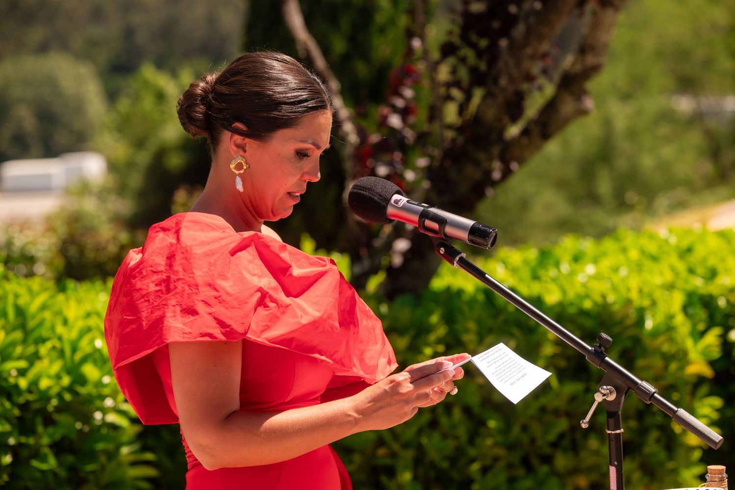 A person in a bright red top speaks into a microphone at an outdoor event with trees in the background.