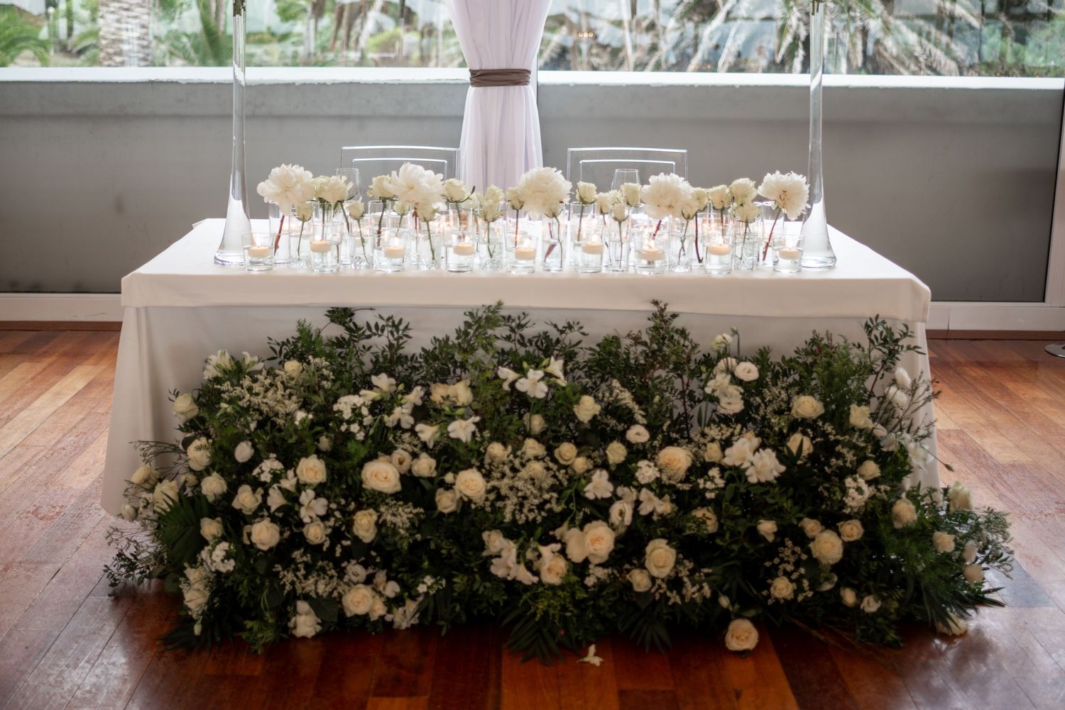 Elegant white floral arrangements adorn tables at a wedding reception with crystal chandeliers overhead.