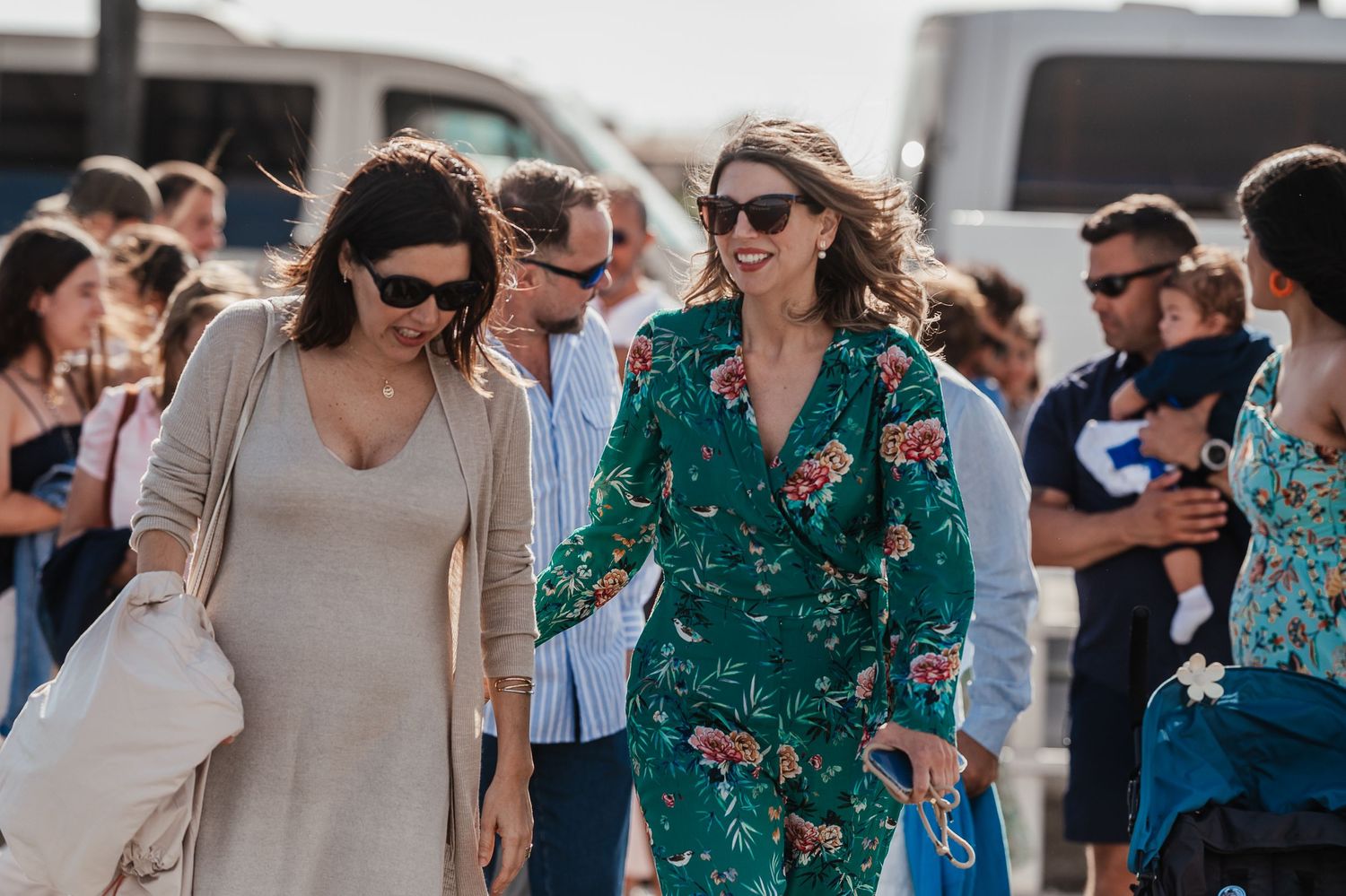 Two friends walk together at an outdoor event, one wearing sunglasses and beige while the other wears a green floral outfit.