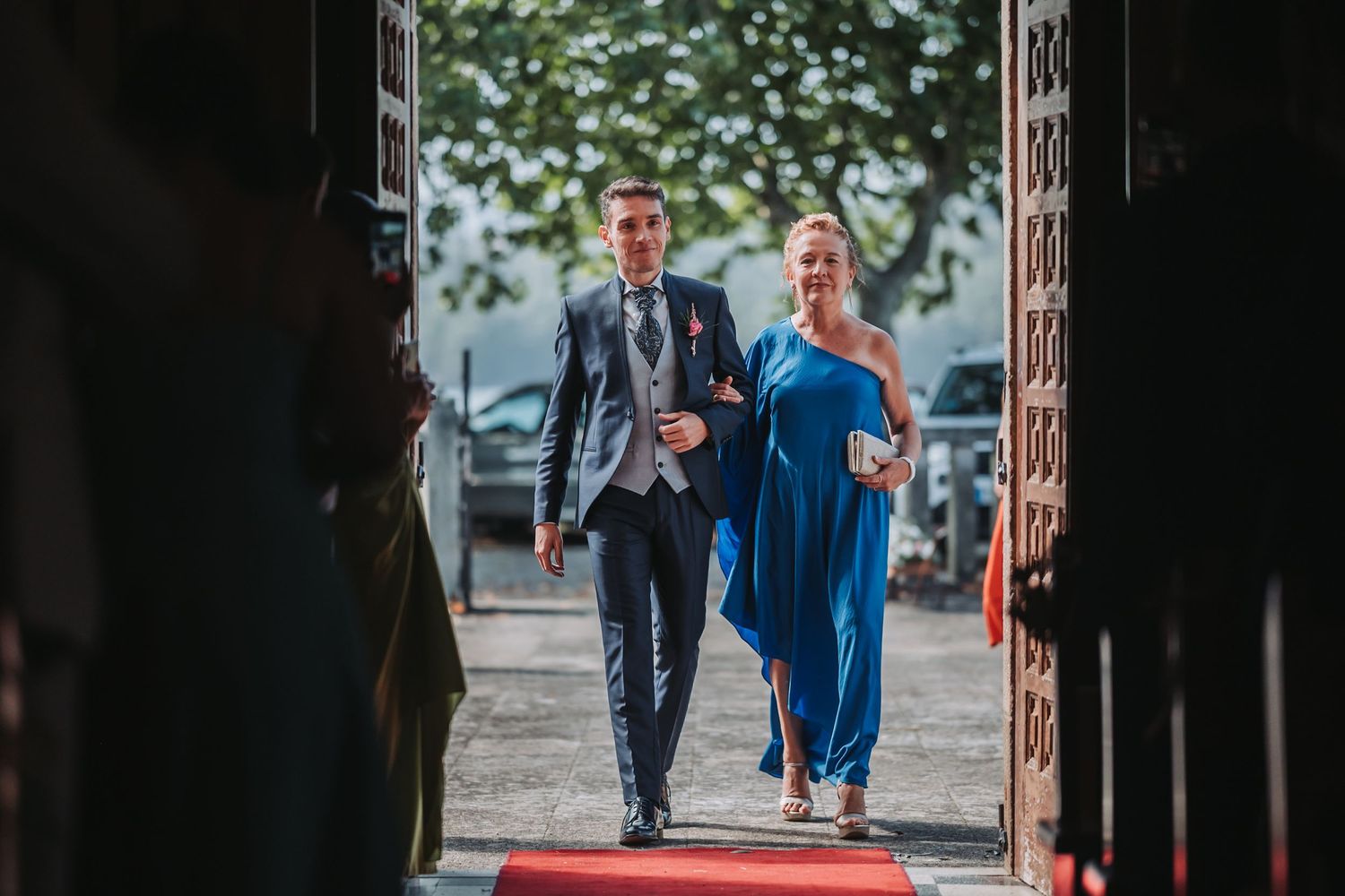 A couple in formal attire walks together through a rustic wooden doorway at an outdoor venue.