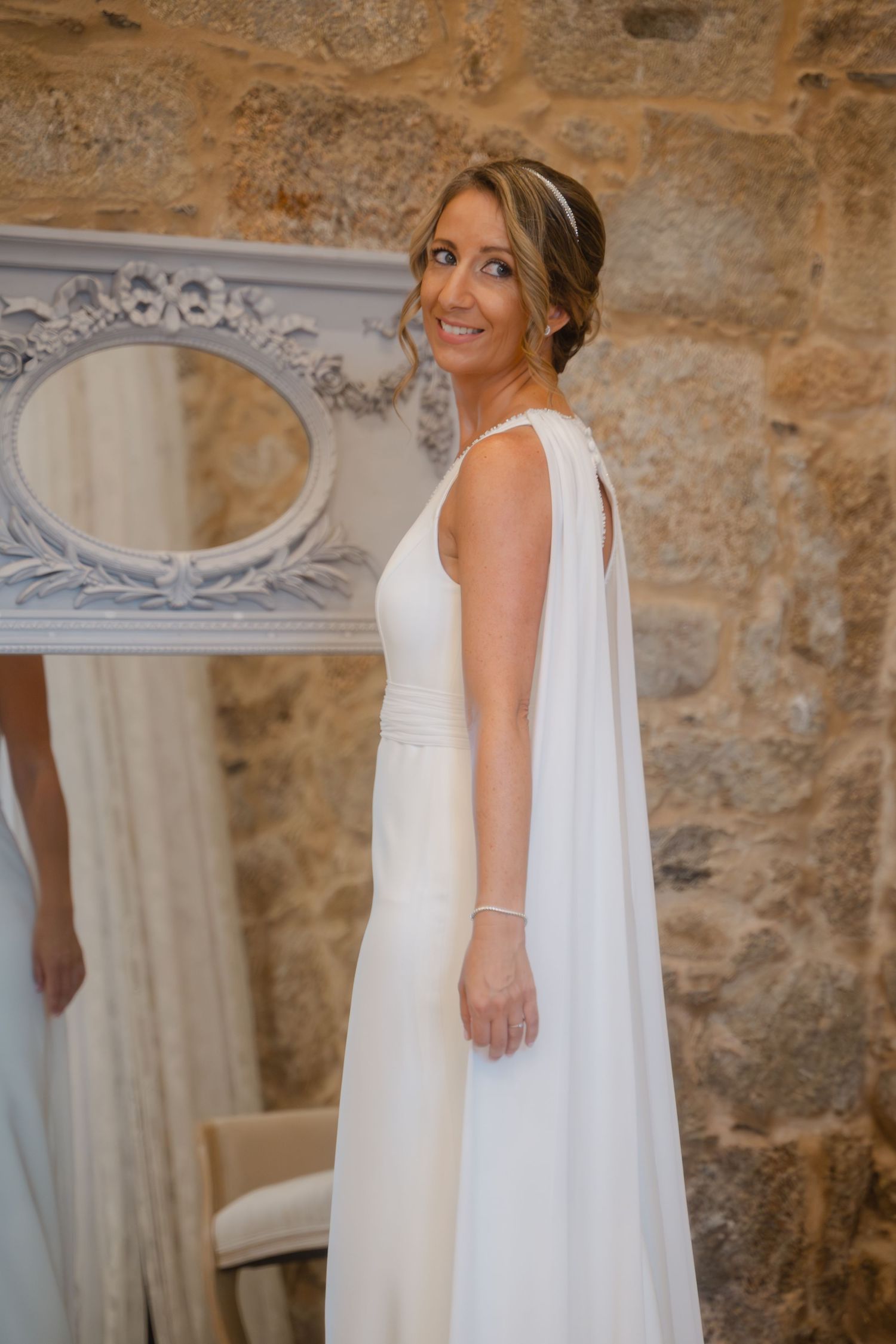 An elegant white wedding dress displayed against a rustic stone wall with ornate mirror details.