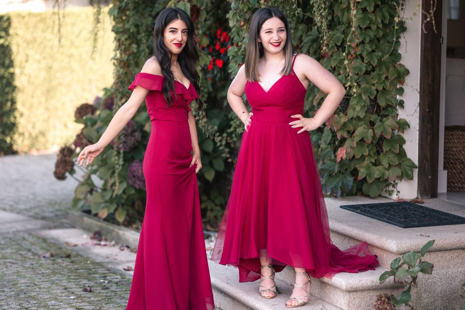 Two people in matching red formal dresses pose together in an outdoor garden setting.