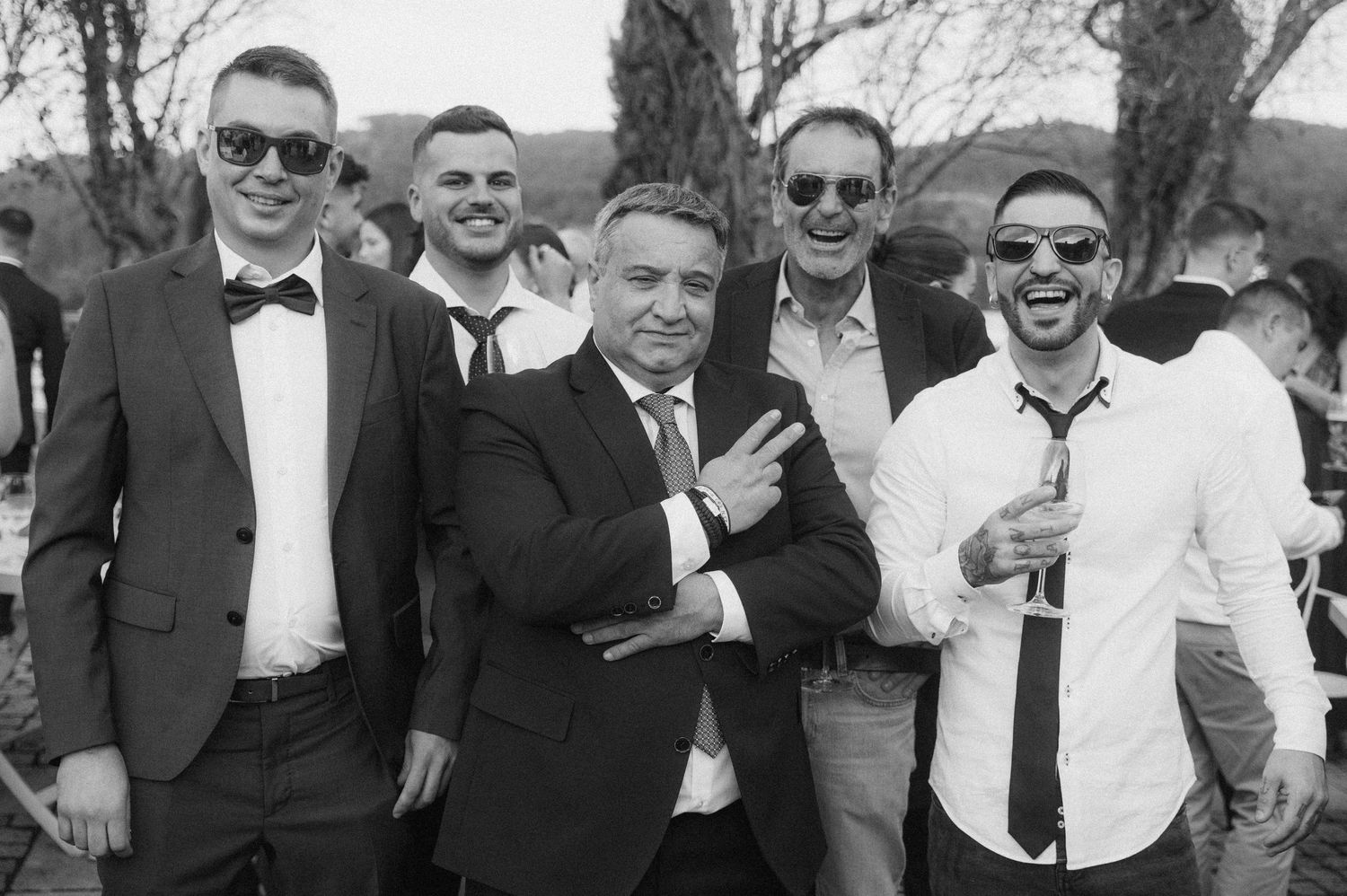 Group of wedding guests in formal attire smiling and posing together outdoors in black and white photo.