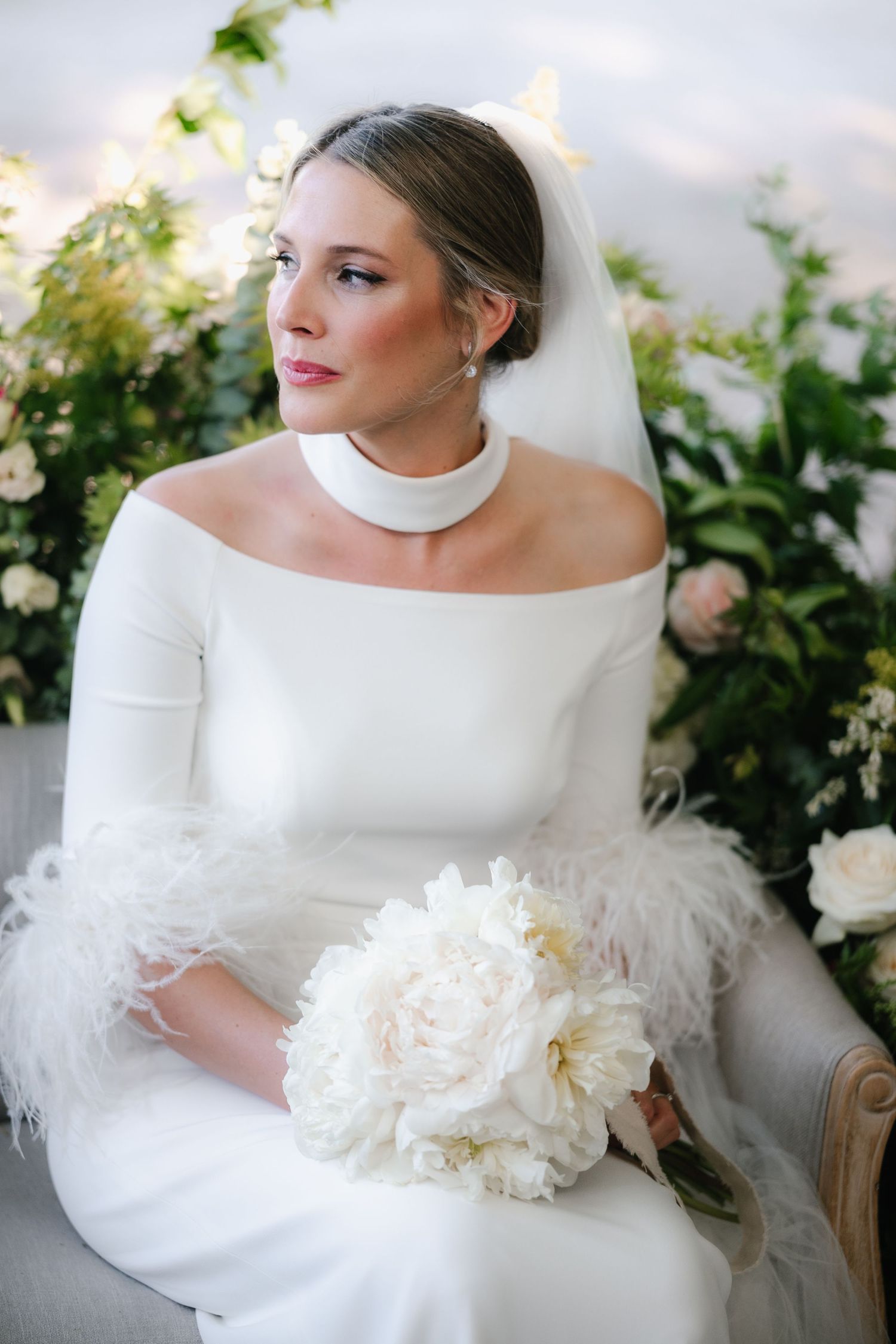 Elegant bride in modern off-shoulder wedding dress with white bouquet poses against floral garden backdrop.