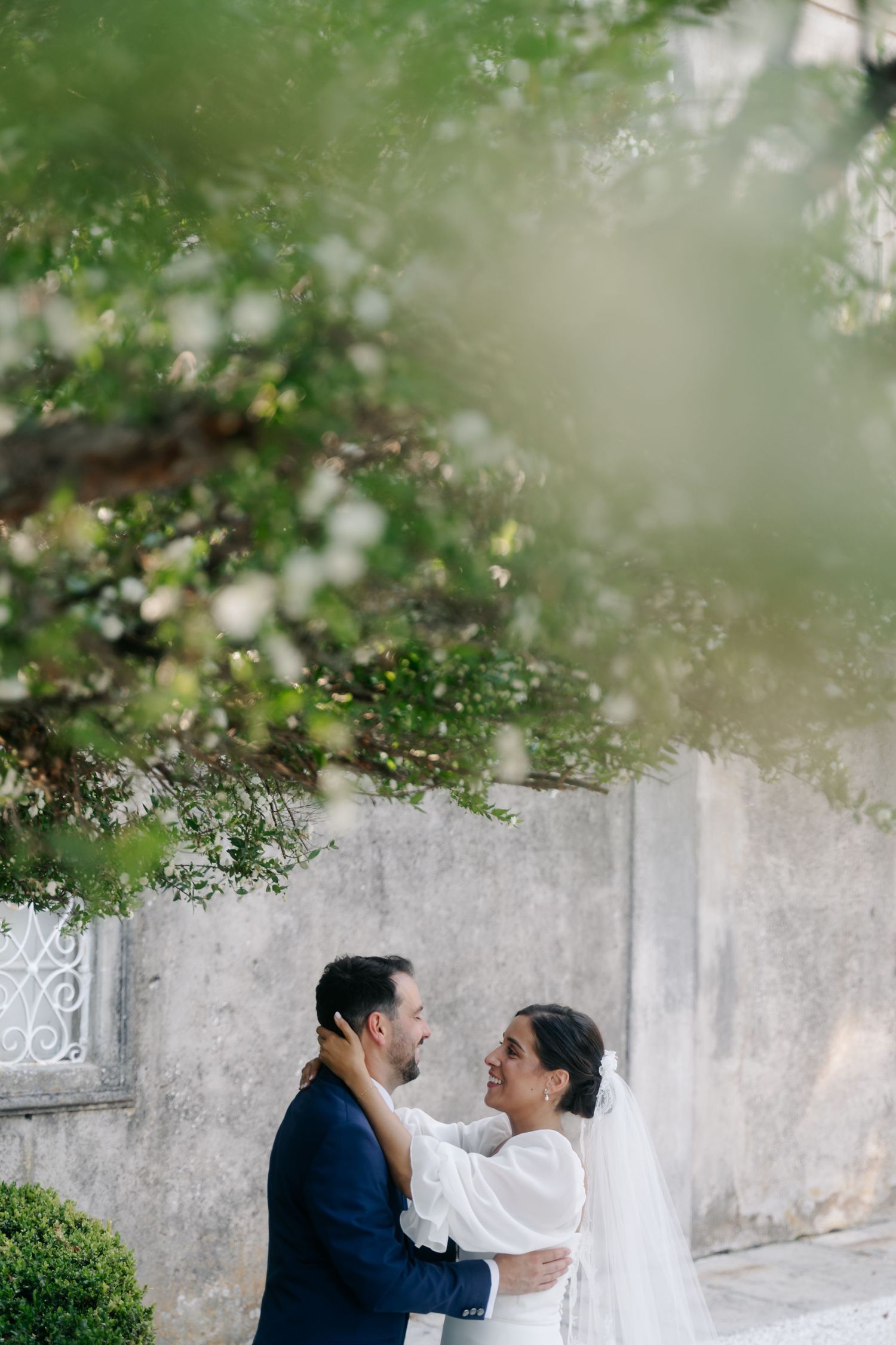 A romantic couple shares an intimate moment against a white wall with lush green foliage overhead in a garden setting.