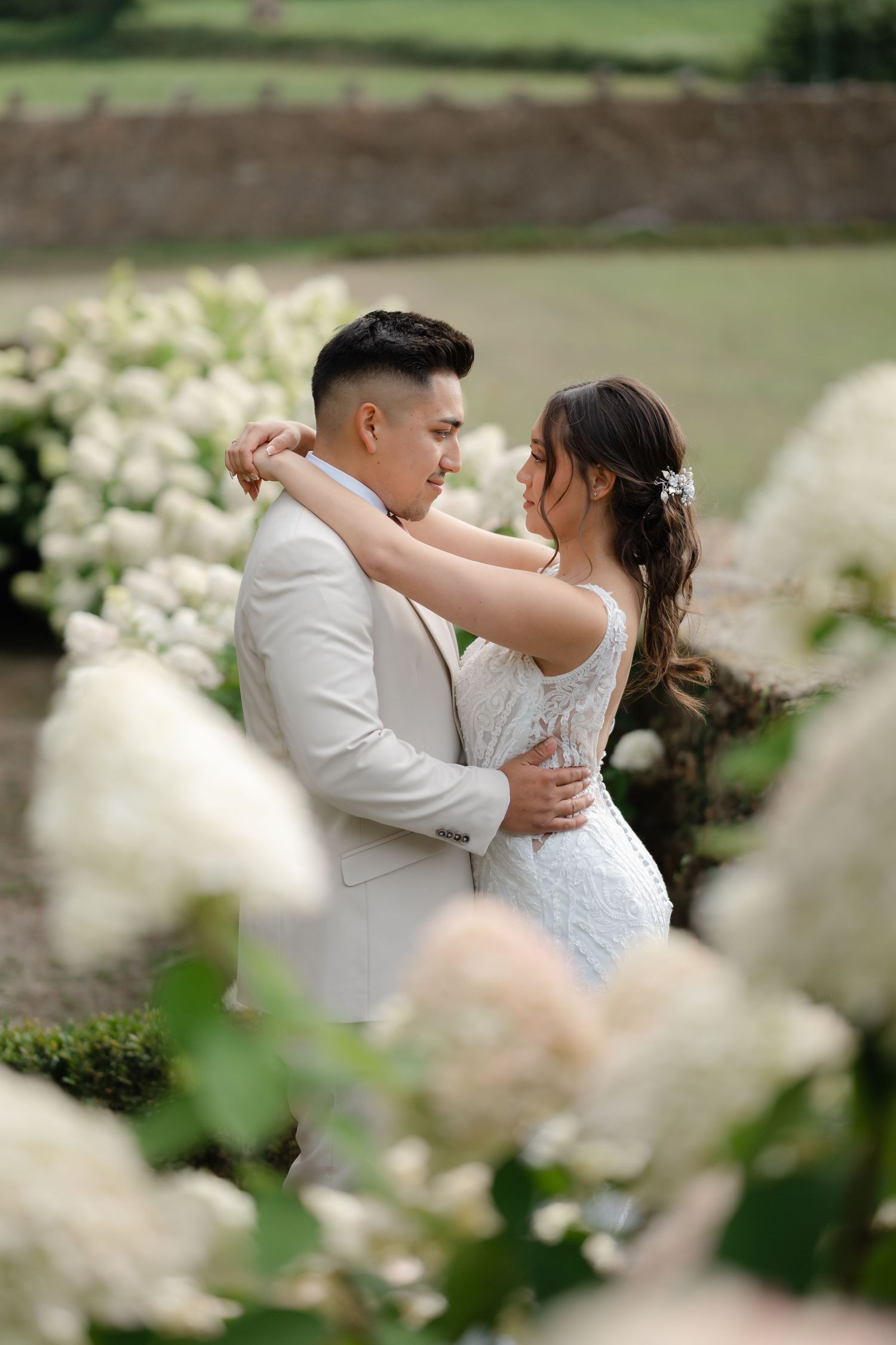 Newlyweds share intimate moments in a garden setting with soft white flowers in the foreground.