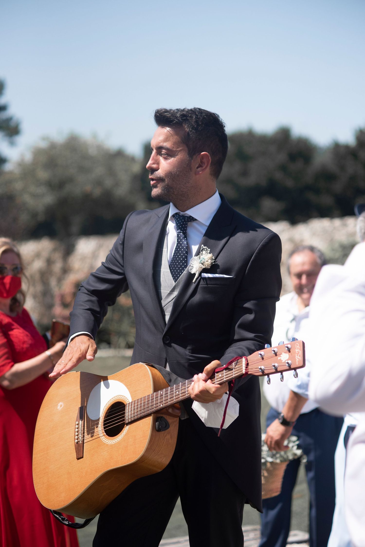 A person in a dark suit plays acoustic guitar outdoors during a wedding ceremony.