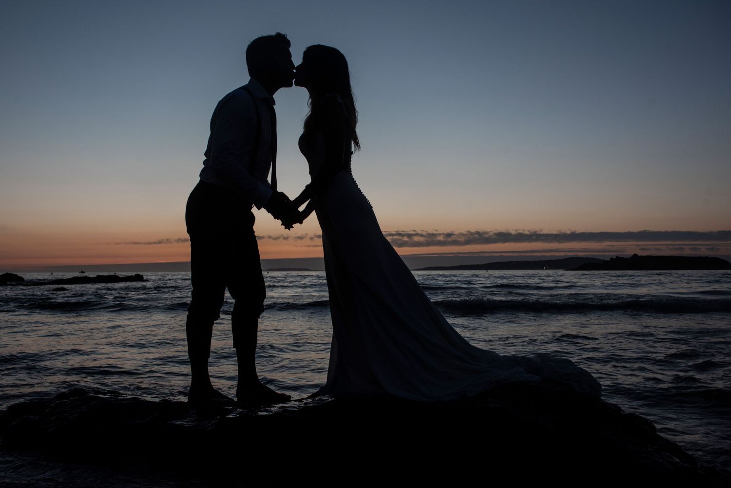 Silhouettes of a couple embrace against dramatic ocean sunset backdrop creating romantic seaside moment.