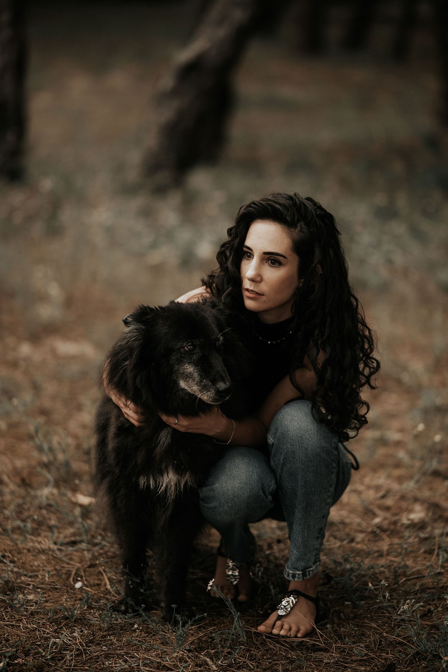 Person in dark clothing crouching on ground with a black dog in a natural outdoor setting.