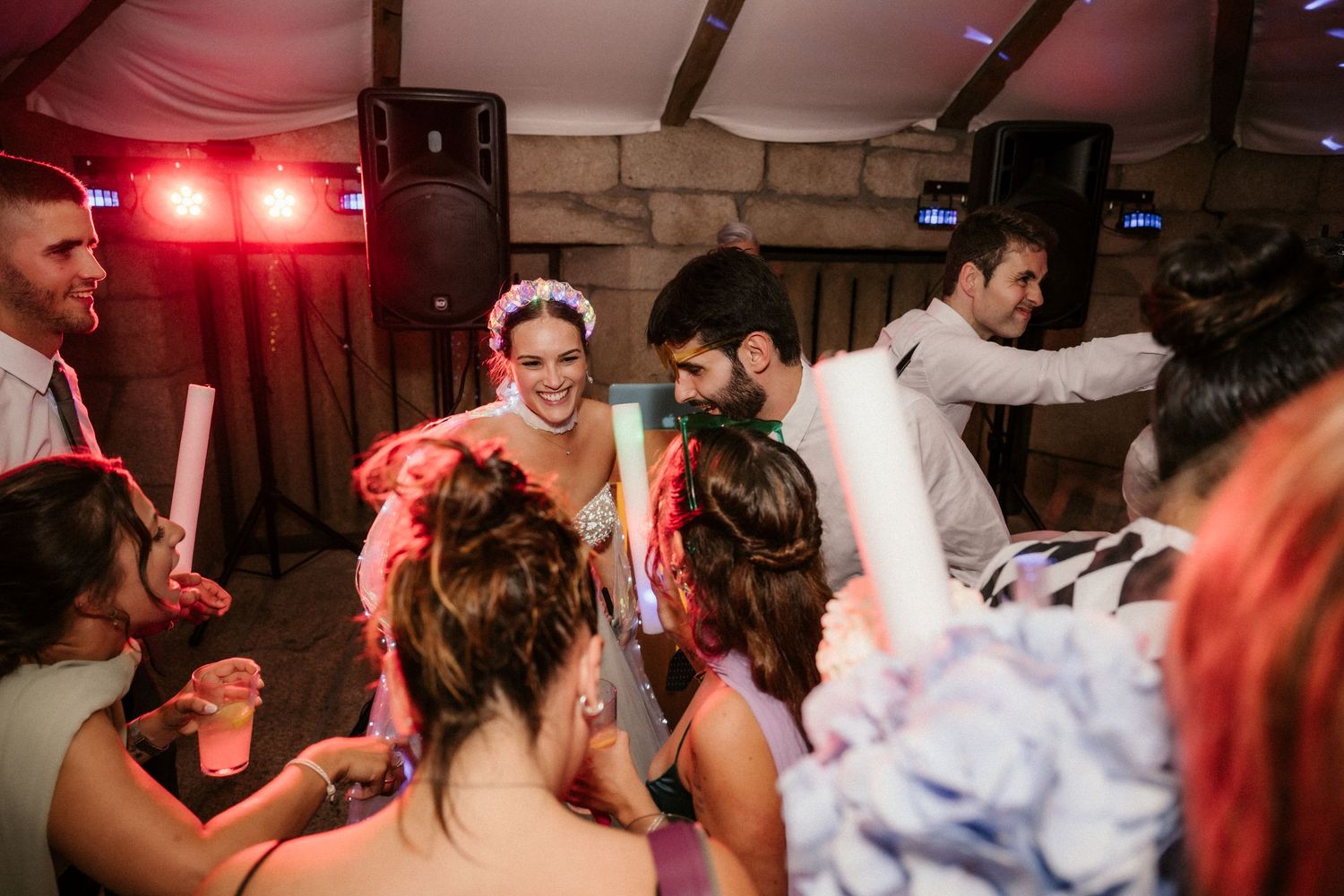 Wedding guests dance and celebrate on a lively dance floor with red lighting in a rustic indoor venue.