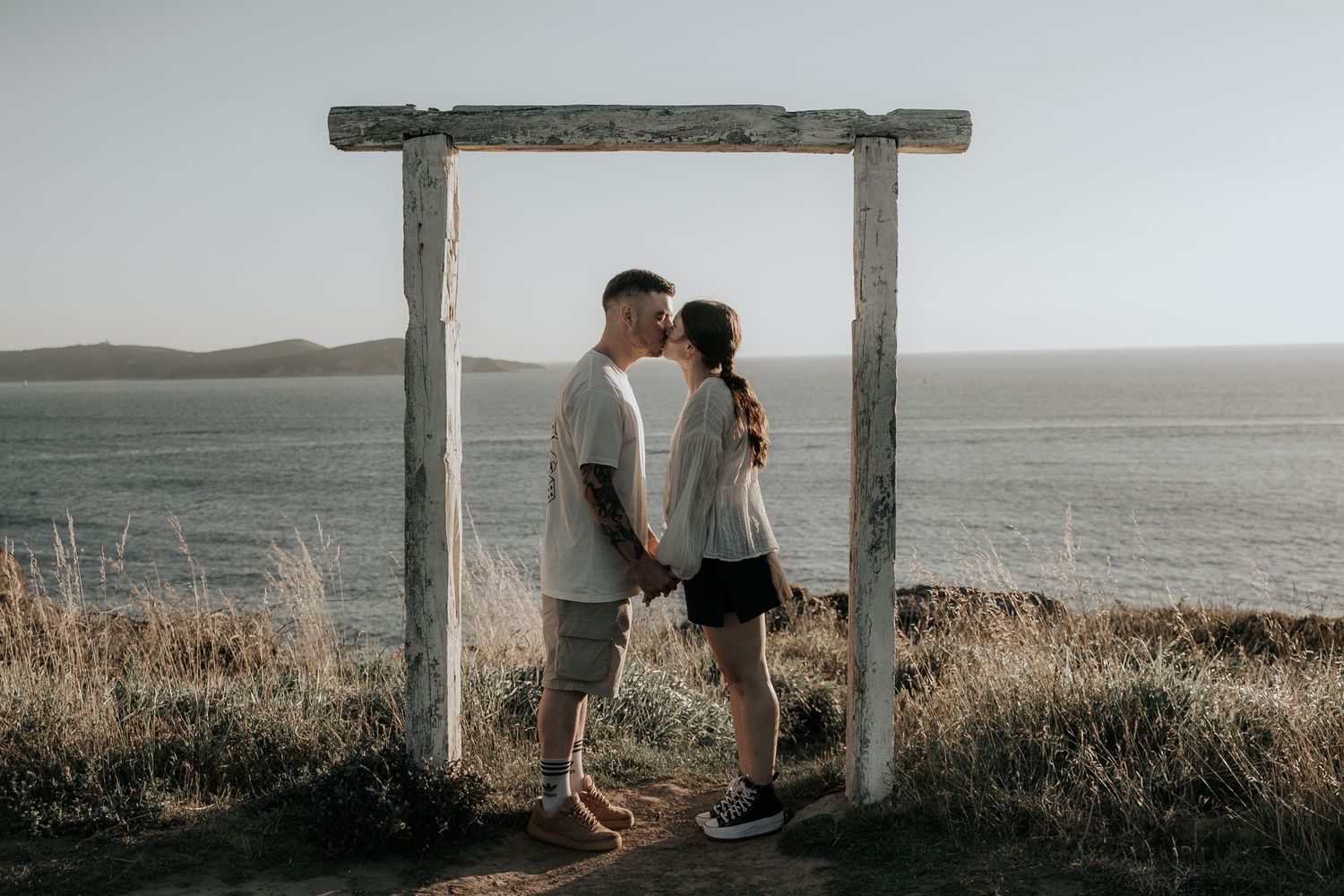 A romantic couple embraces under a rustic wooden archway overlooking the ocean.