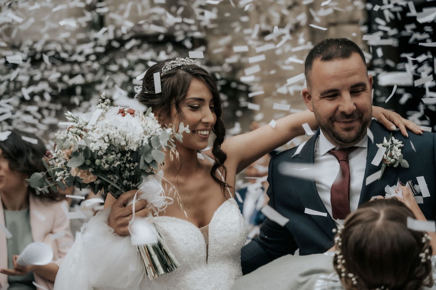Newlyweds celebrate with confetti during outdoor wedding ceremony while holding white floral bouquet.