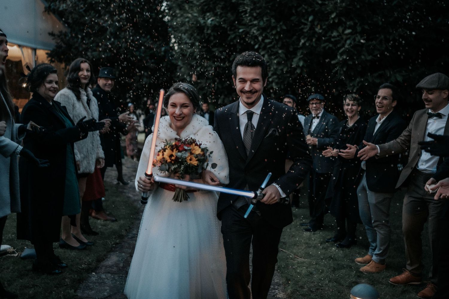Wedding guests form an arch with lightsabers as a couple walks beneath them at an outdoor evening celebration.