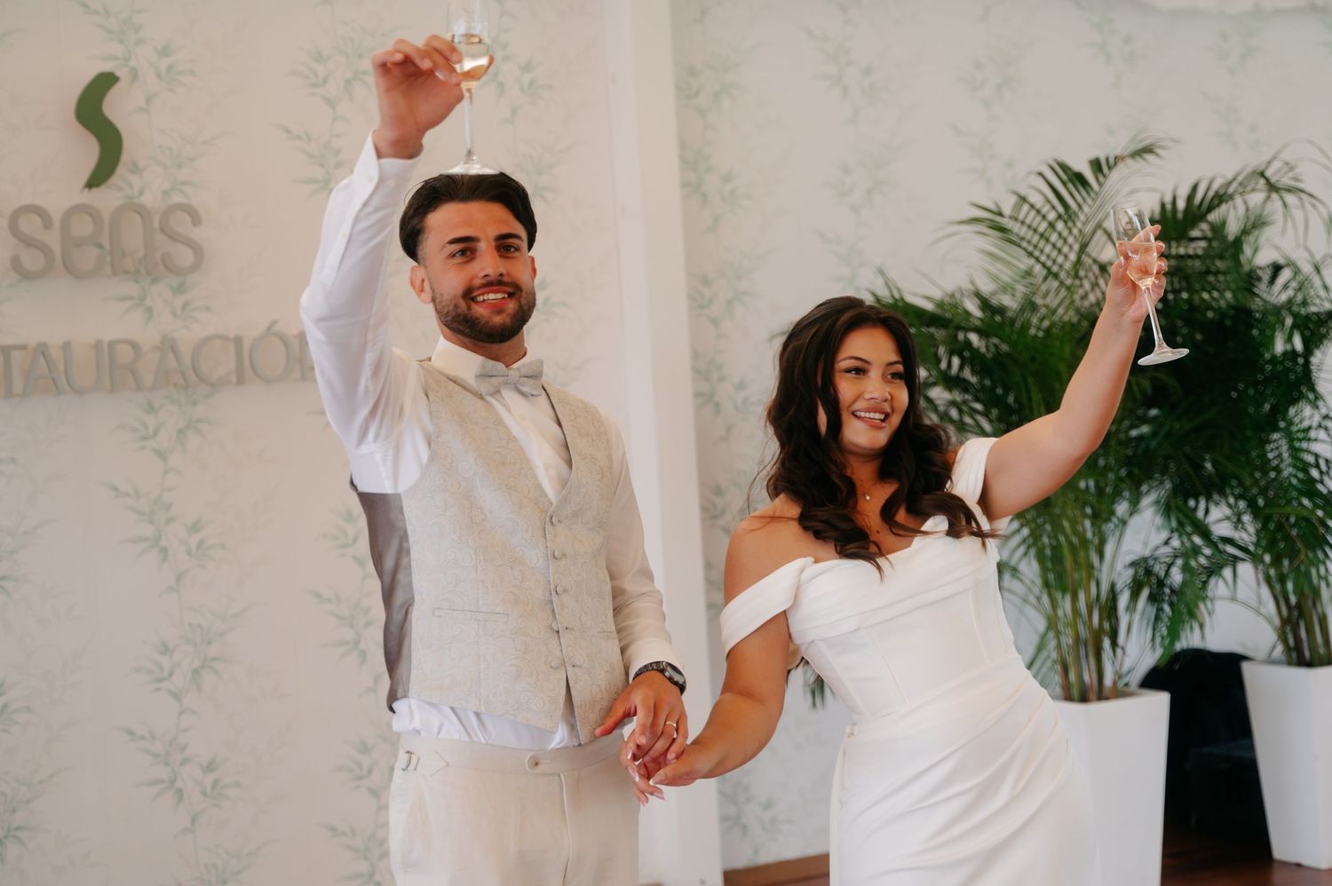 A couple in white outfits dance together in a bright indoor space with tropical plants in the background.
