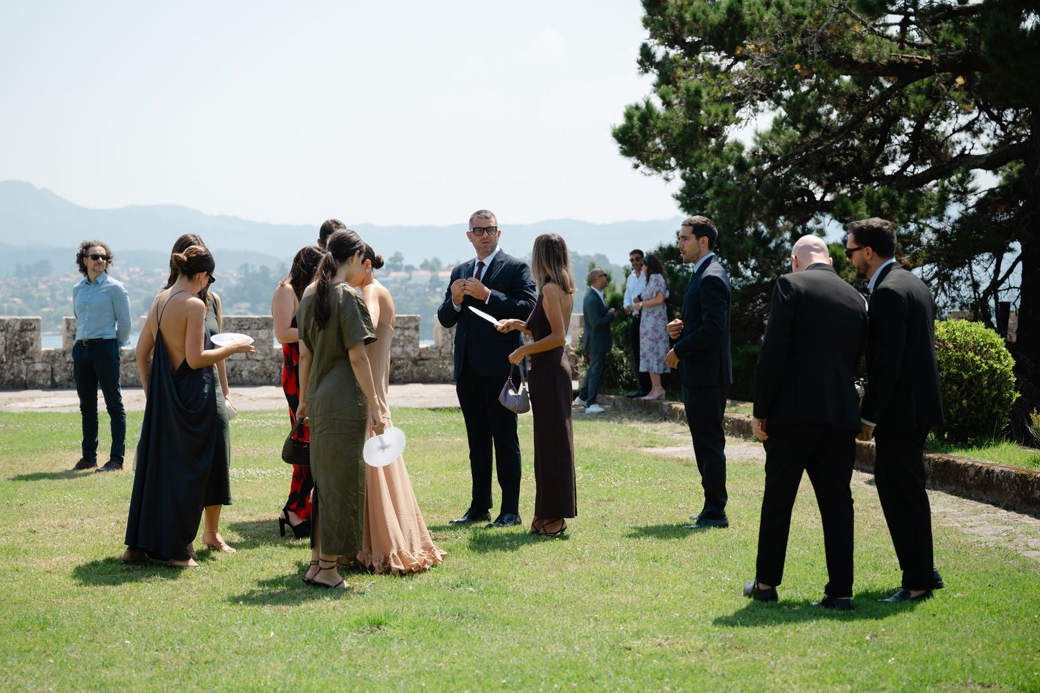A small outdoor wedding ceremony takes place on a grassy area with guests gathered in a semi-circle formation.