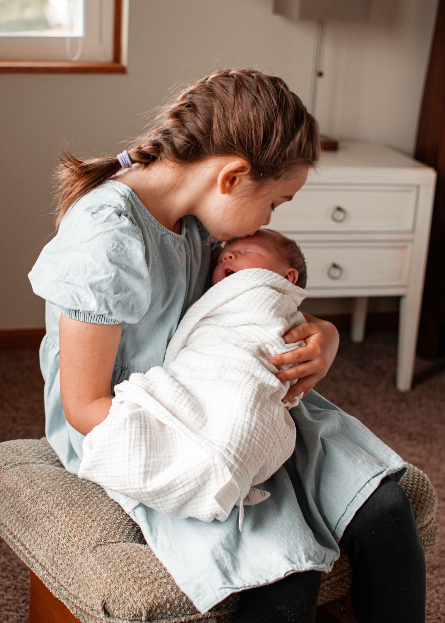 Intimate family photography capturing sweet sibling bond as young girl gently snuggles newborn baby in soft window light